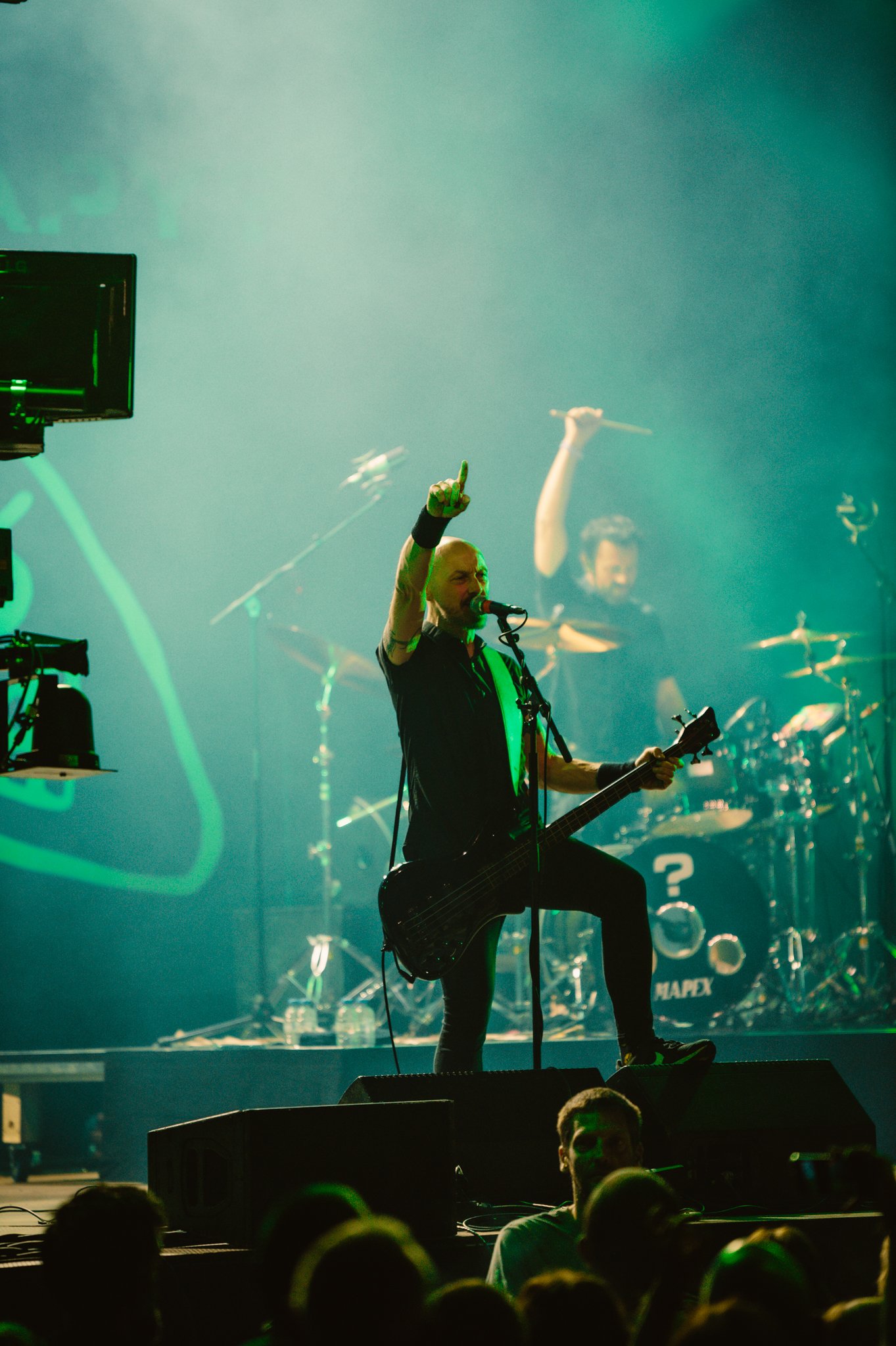 Live concert scene with a guitarist and a drummer on stage, performing energetically in front of a crowd under green lighting.