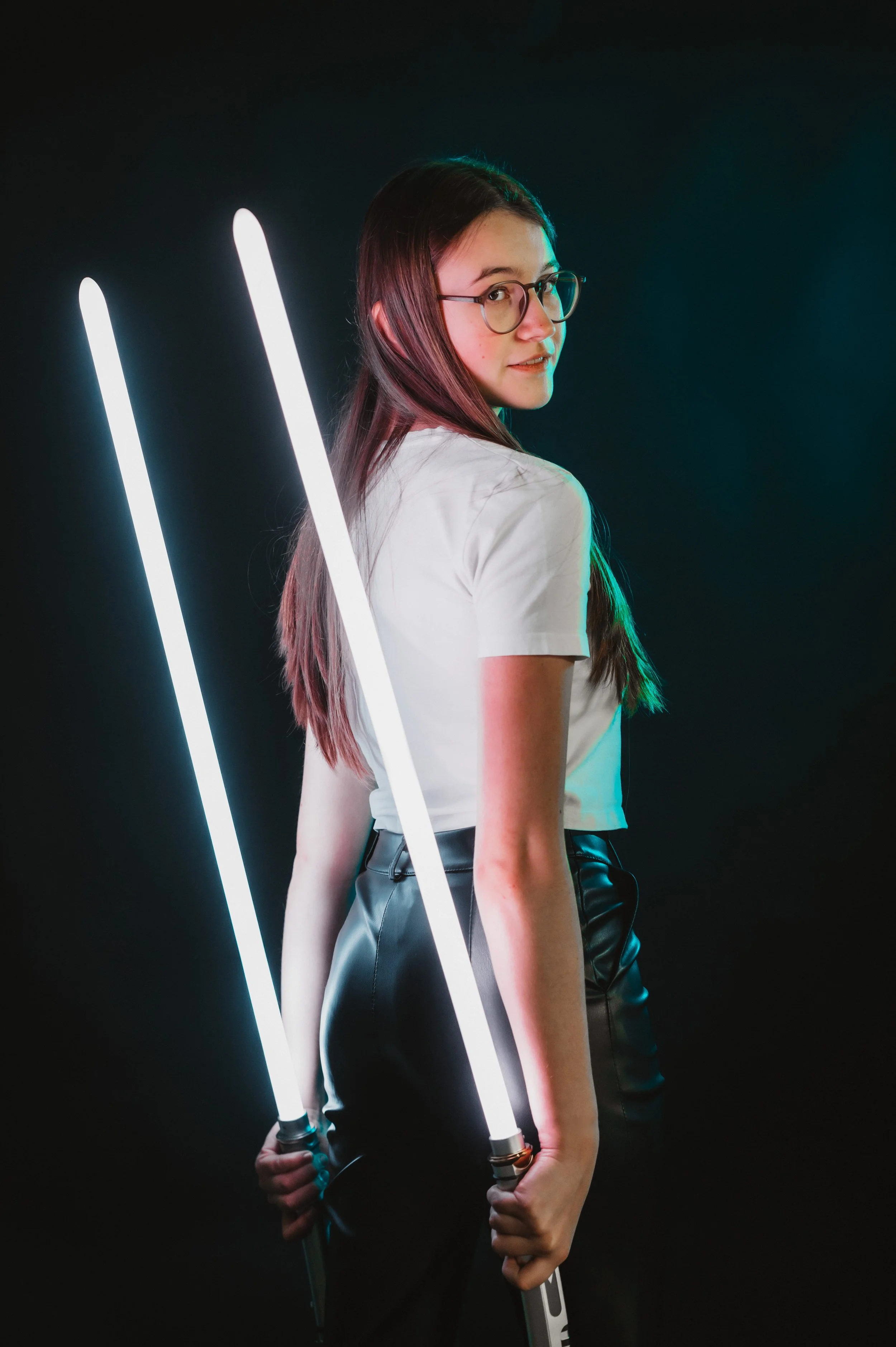 Person wearing glasses and holding two light sabers, standing against a dark background.