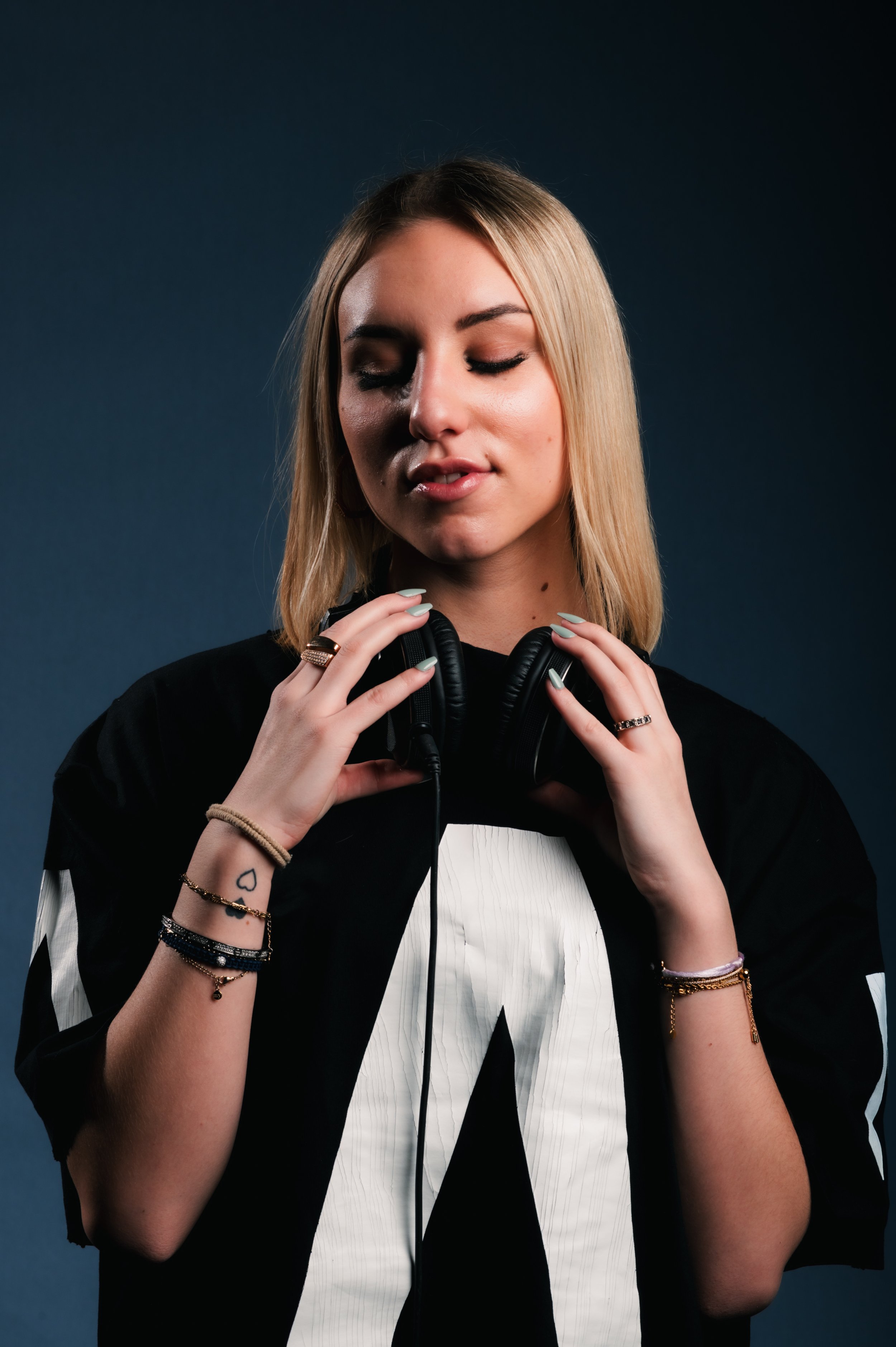 Woman with headphones, wearing a black shirt with a large white letter, eyes closed, blonde hair, and bracelets on both wrists, against a dark background.