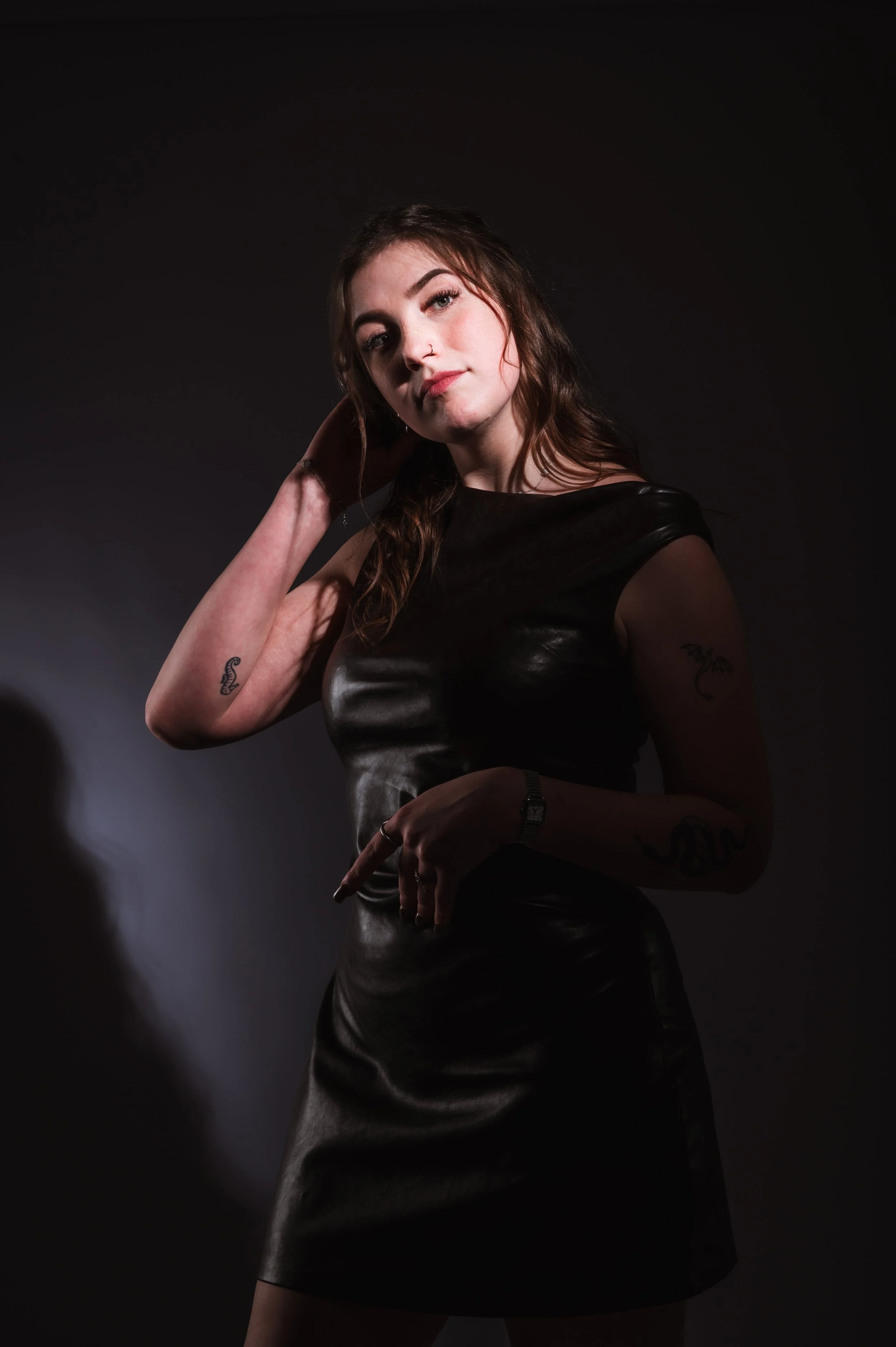 Young woman with tattoos, wearing a black leather dress, posing against a dark background.