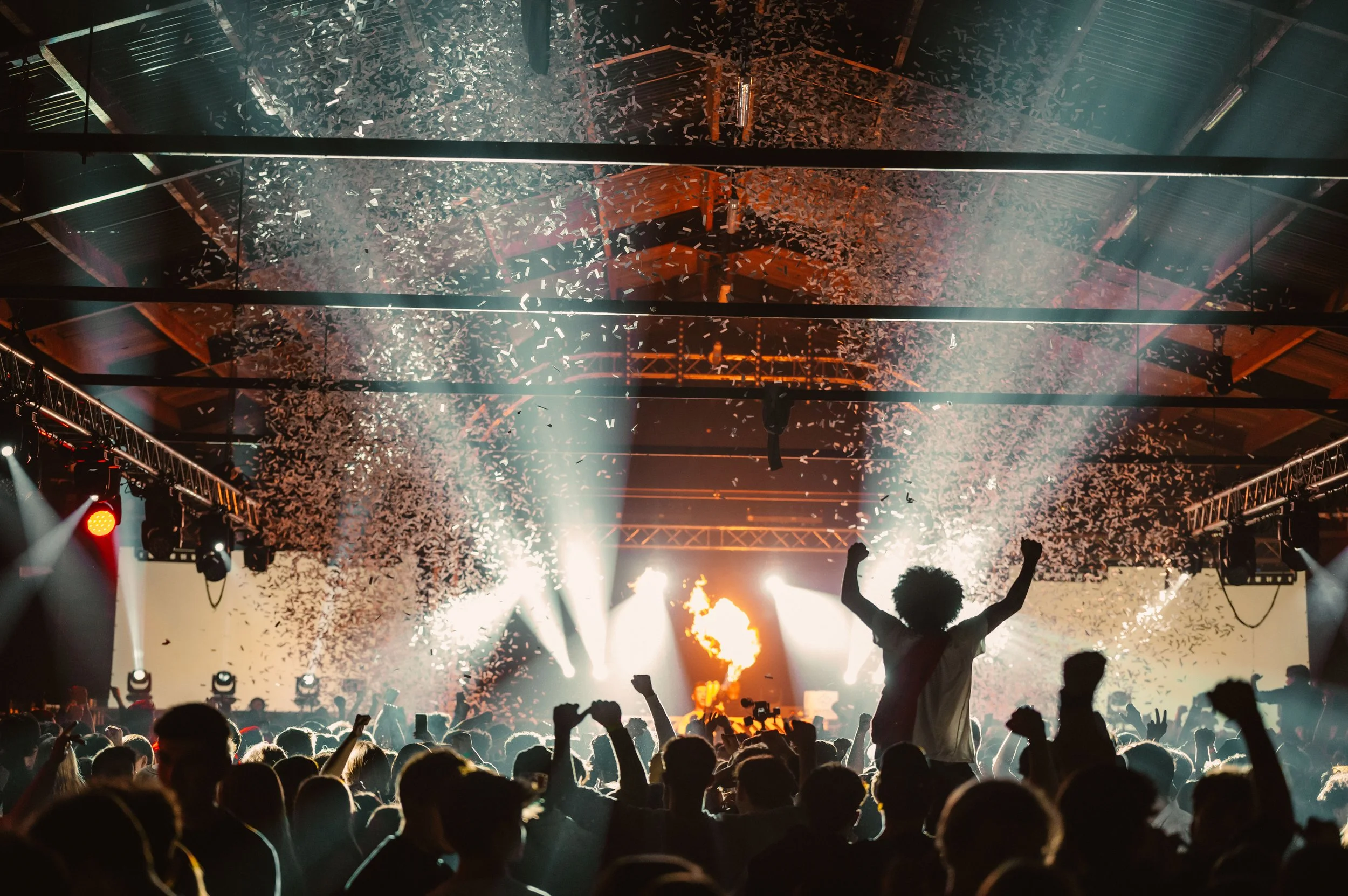 Concert audience with raised arms, confetti falling, colorful stage lights.