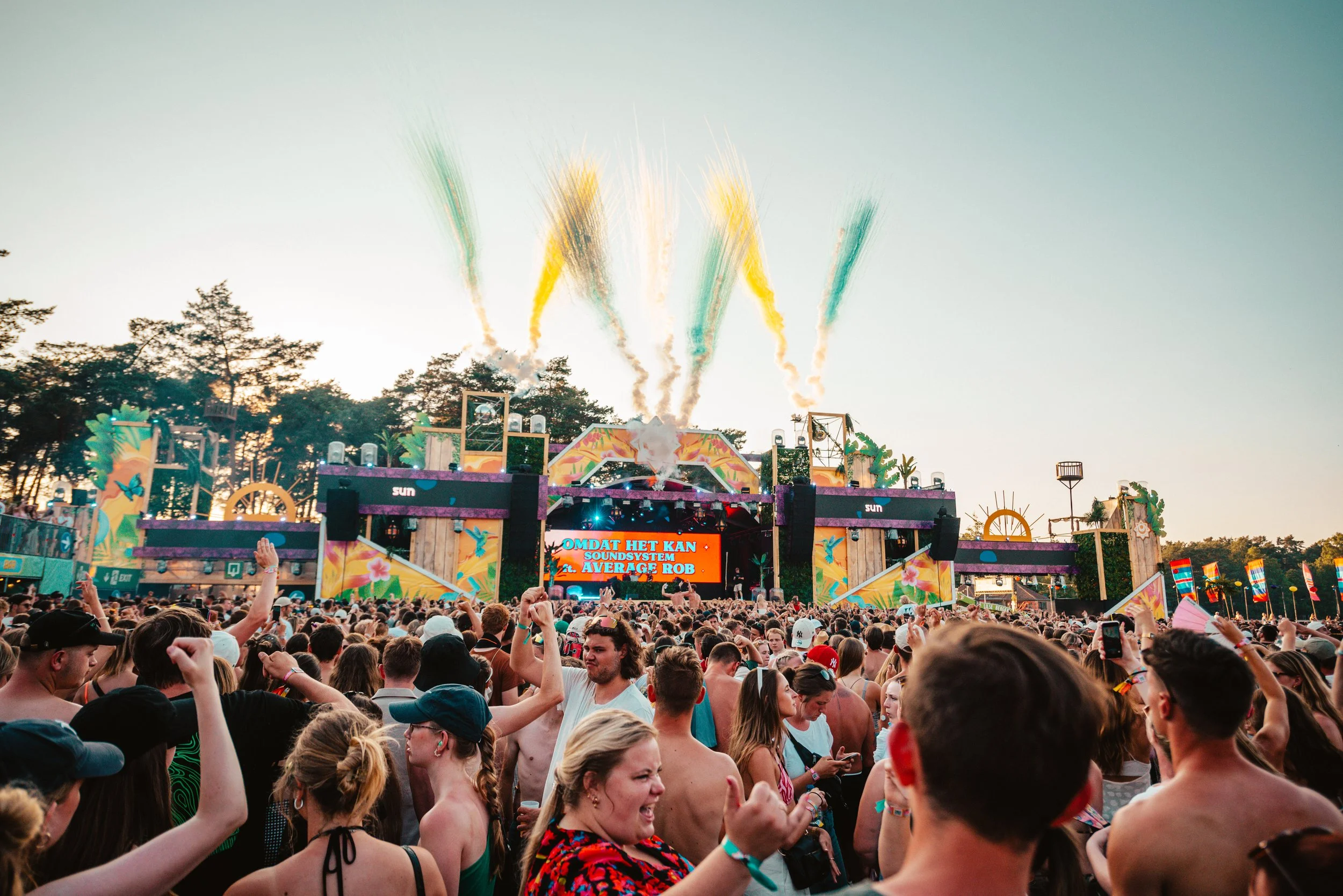 Outdoor music festival with a large crowd in front of a stage, colorful fireworks in the sky, and vibrant decorations.