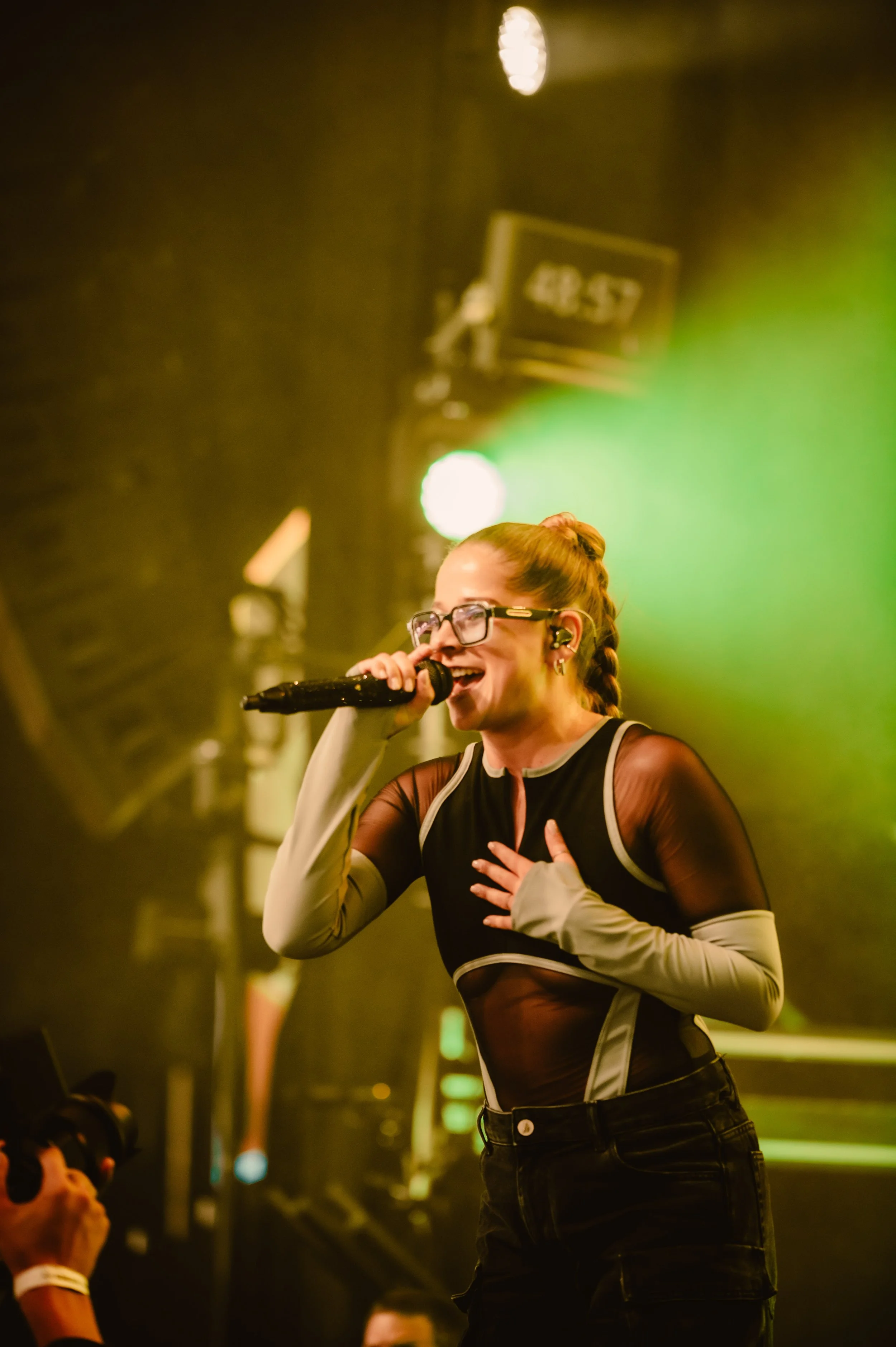 Singer with glasses and braid performing on stage with microphone and bright lighting.