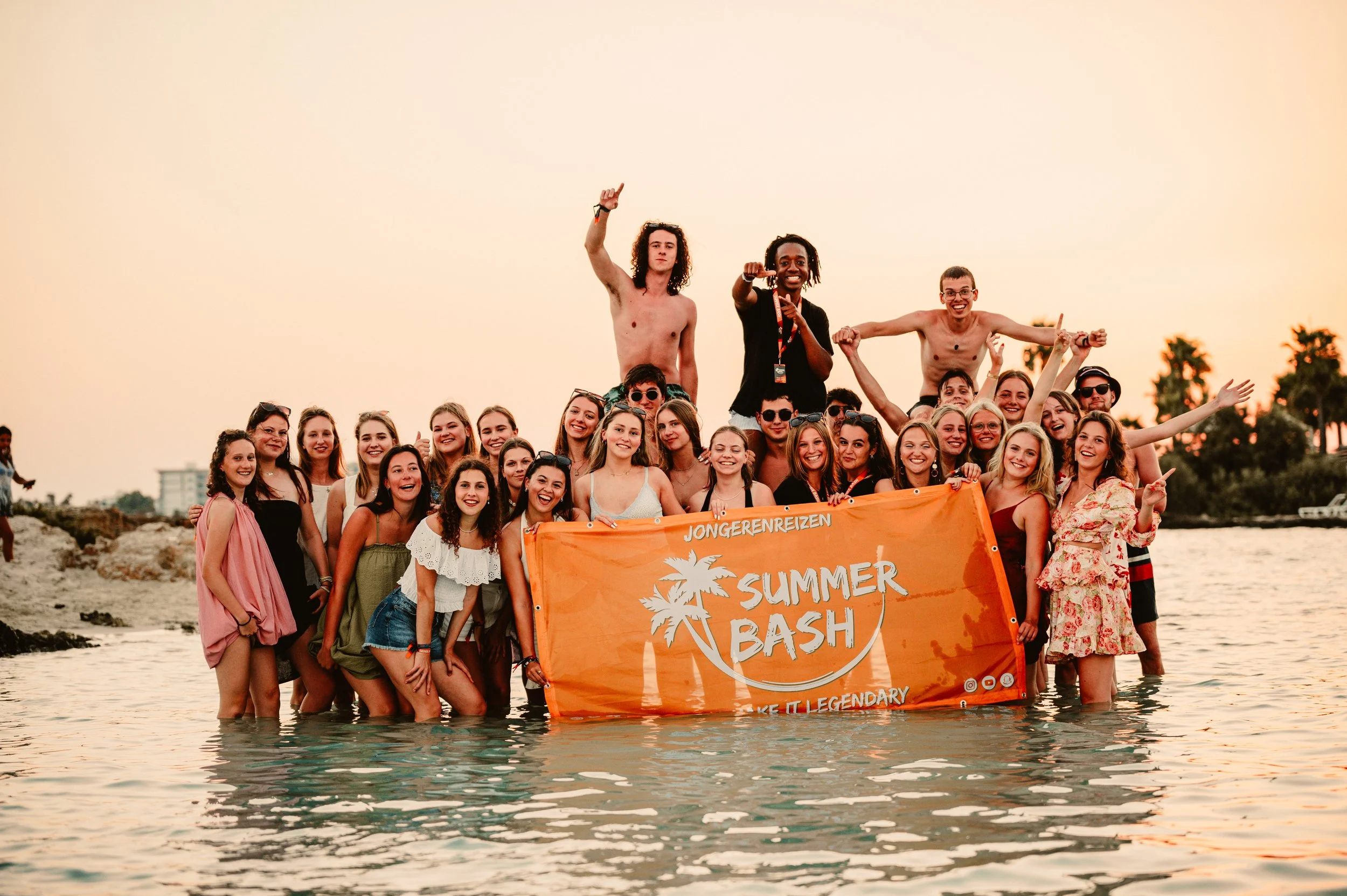 A group of people smiling and posing in shallow water, holding an orange banner that reads "Summer Bash" with palm tree graphics. The photo is set during sunset with palm trees in the background.