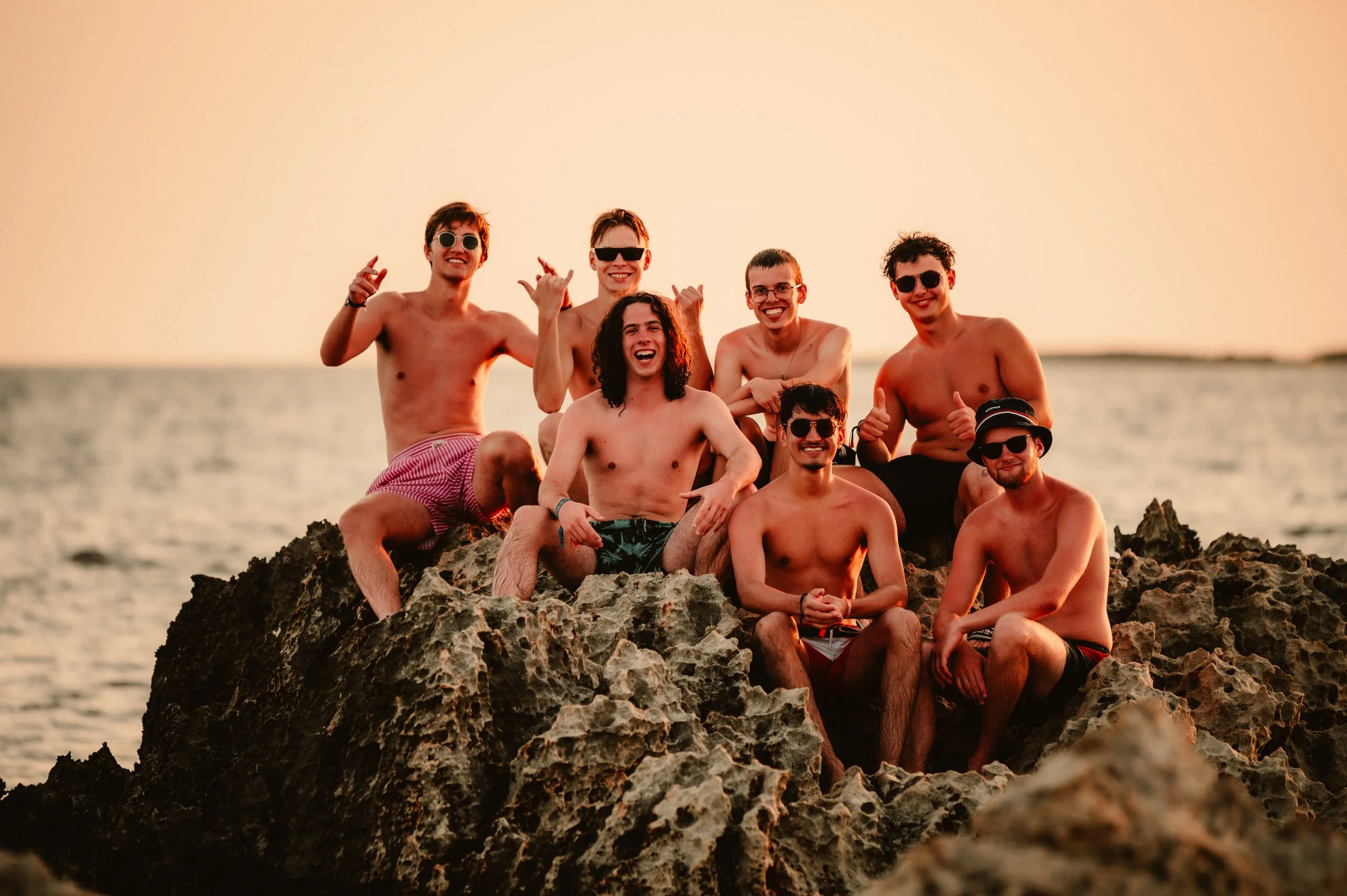 Group of seven young men posing shirtless on rocky shoreline near the ocean during sunset.