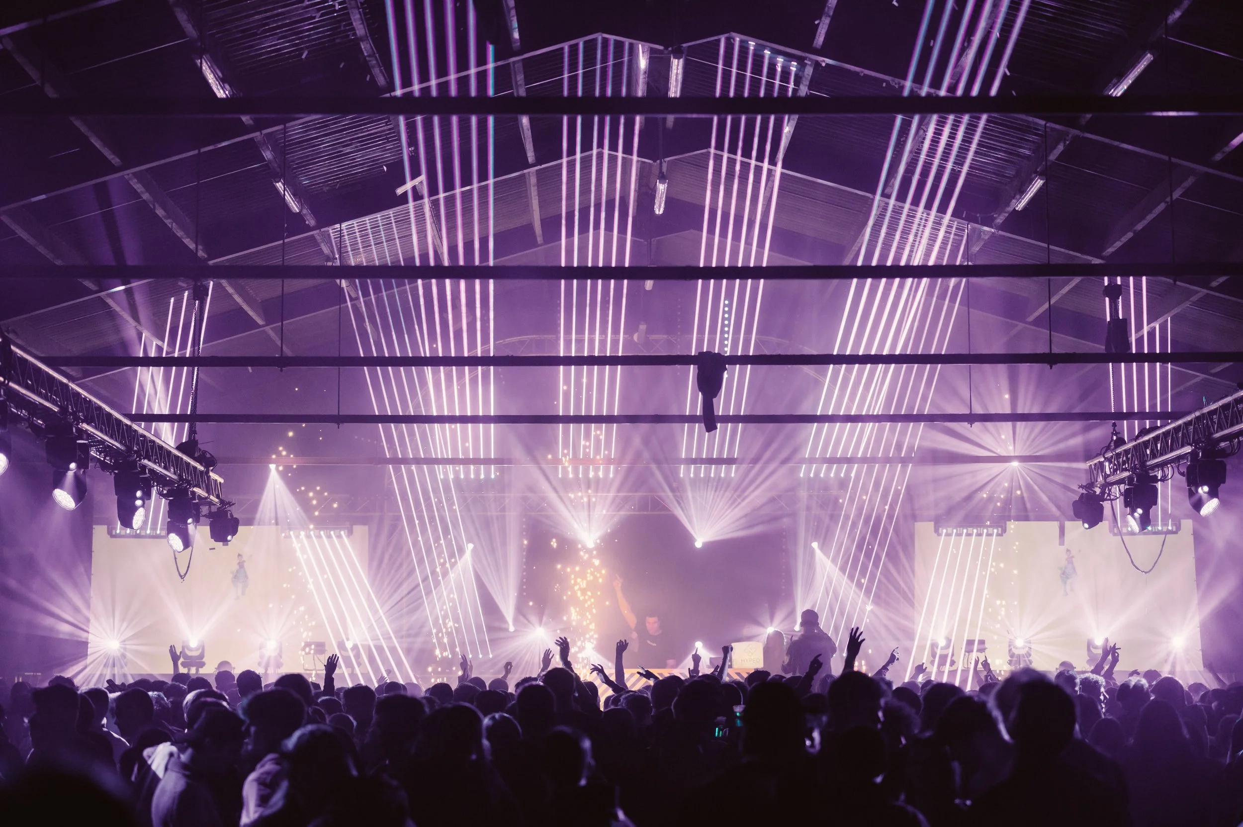 Crowded concert venue with purple lighting and audience raising hands, stage with DJ or performer, and dynamic light beams.