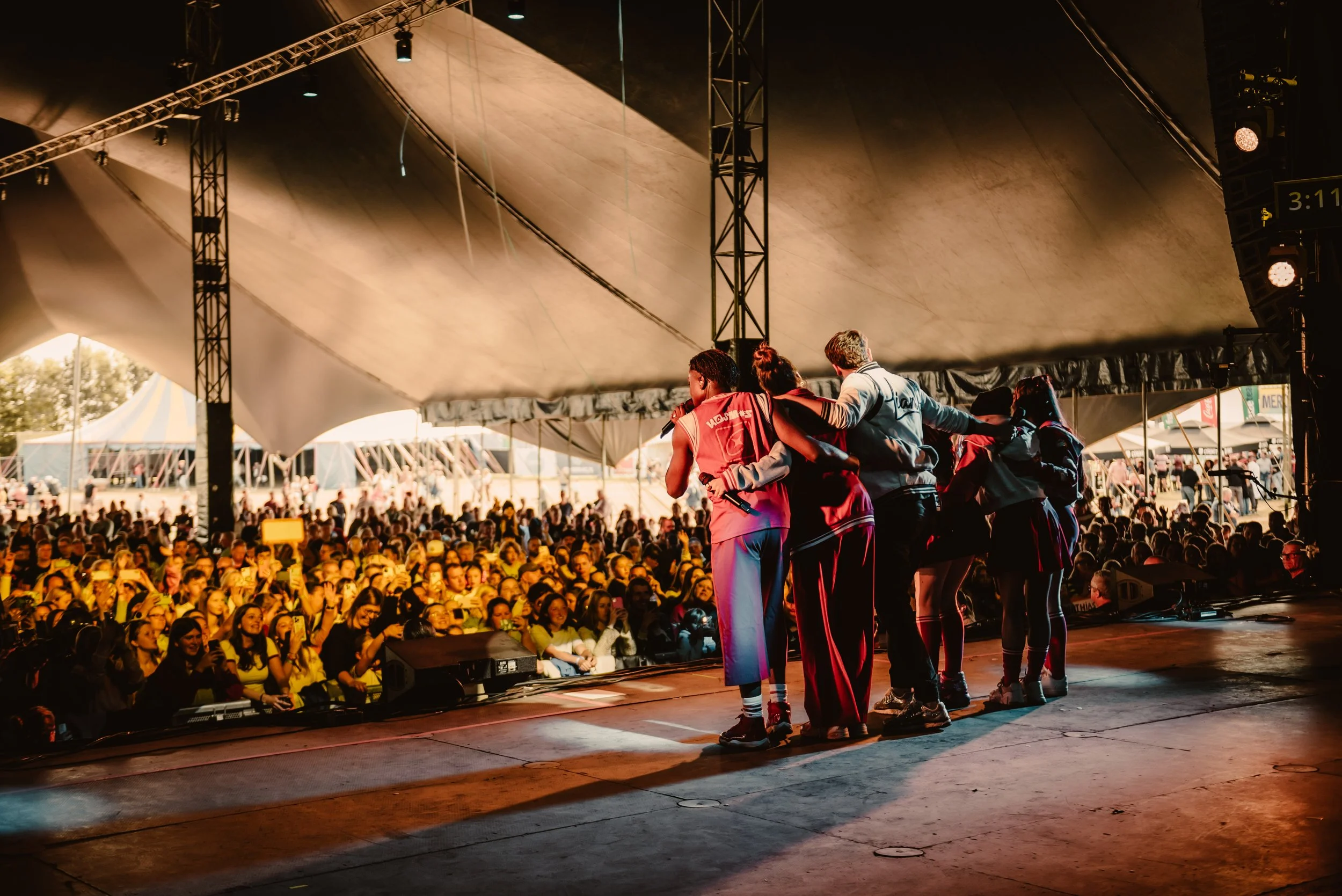People on stage at a large outdoor event, facing a cheering crowd, under a tent canopy.