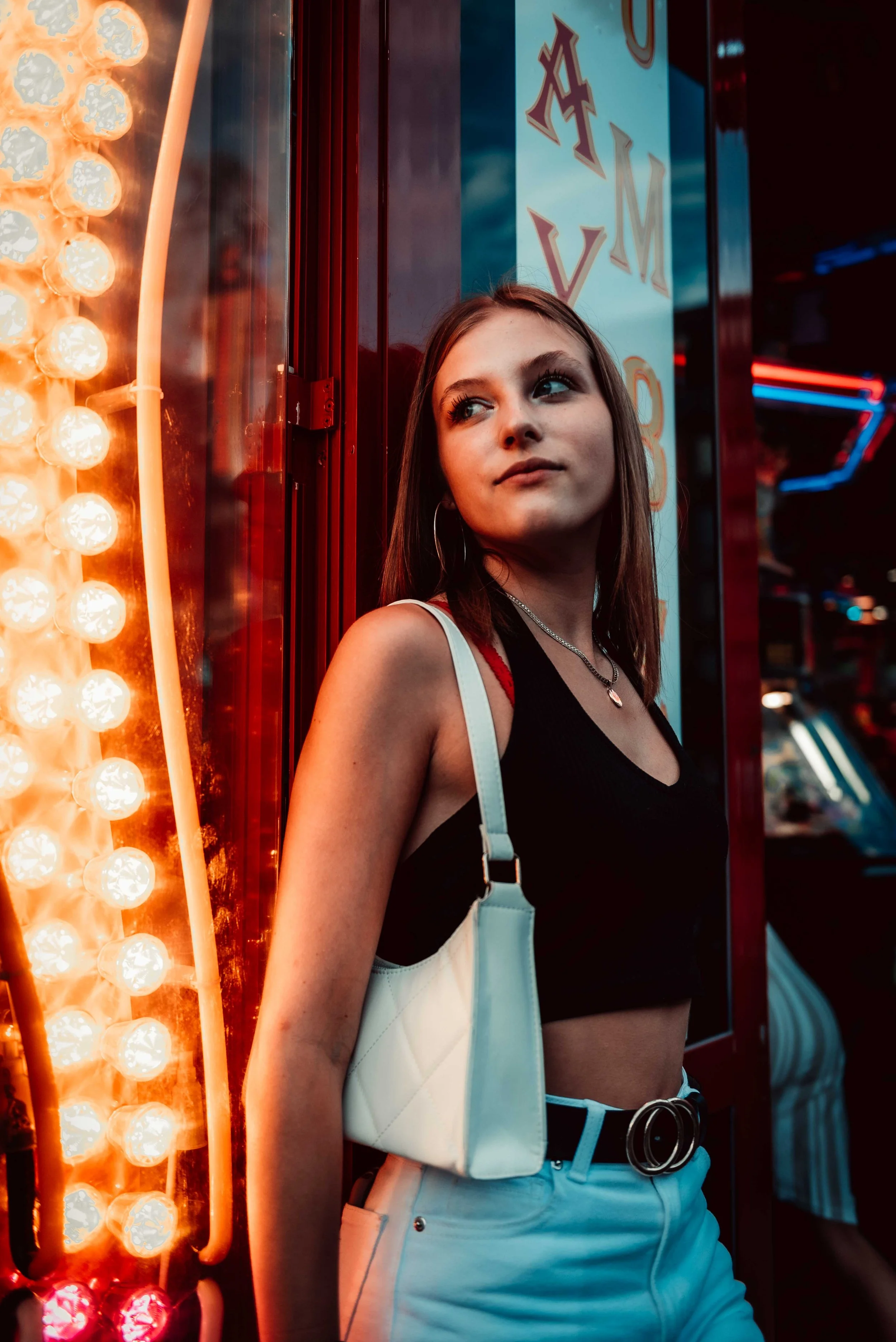 Young woman leaning against a lit wall with neon lights, wearing a black crop top, white pants, and holding a white purse.