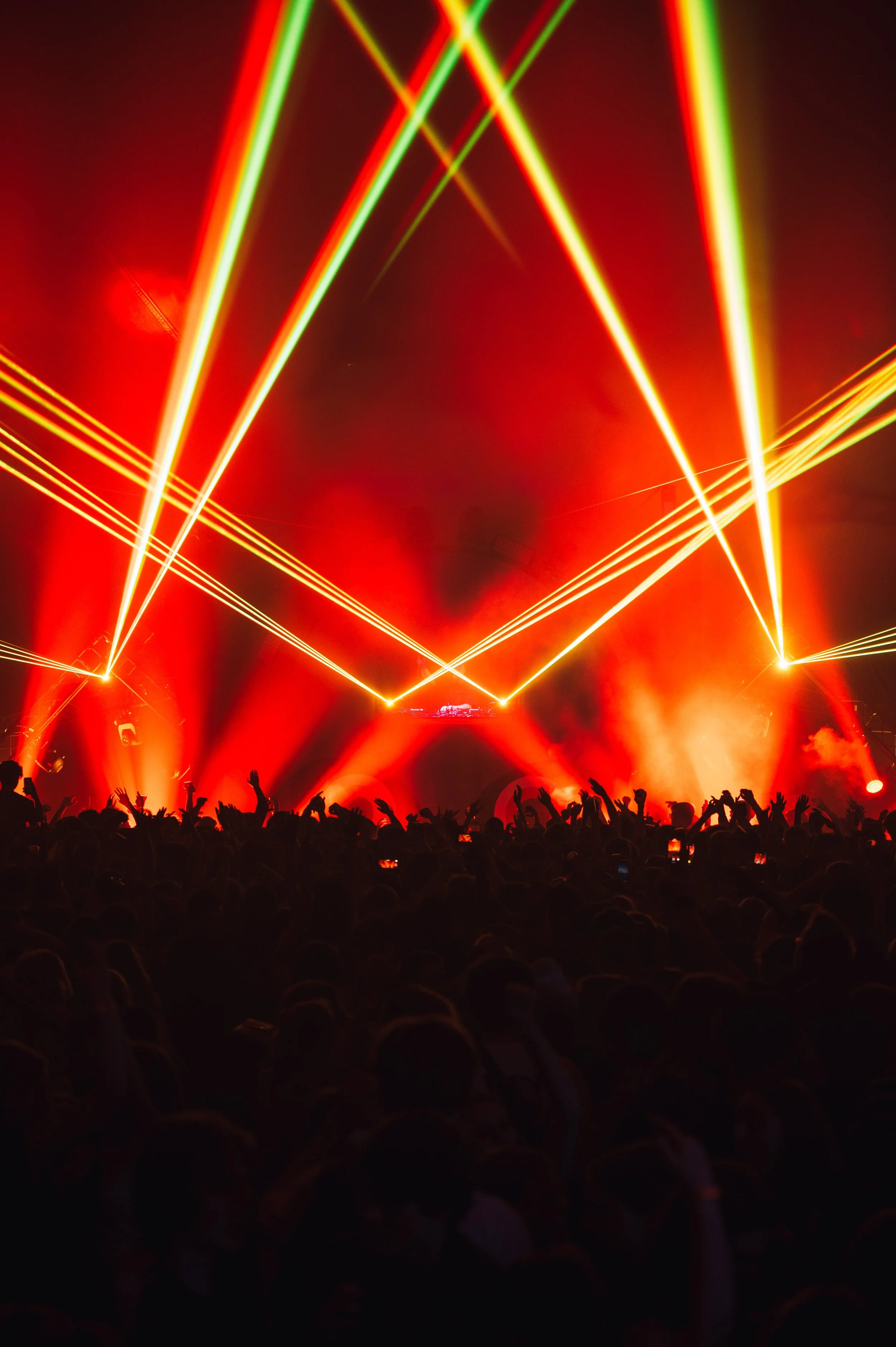 Crowd enjoying a concert with red and yellow laser lights and a DJ booth in the background.