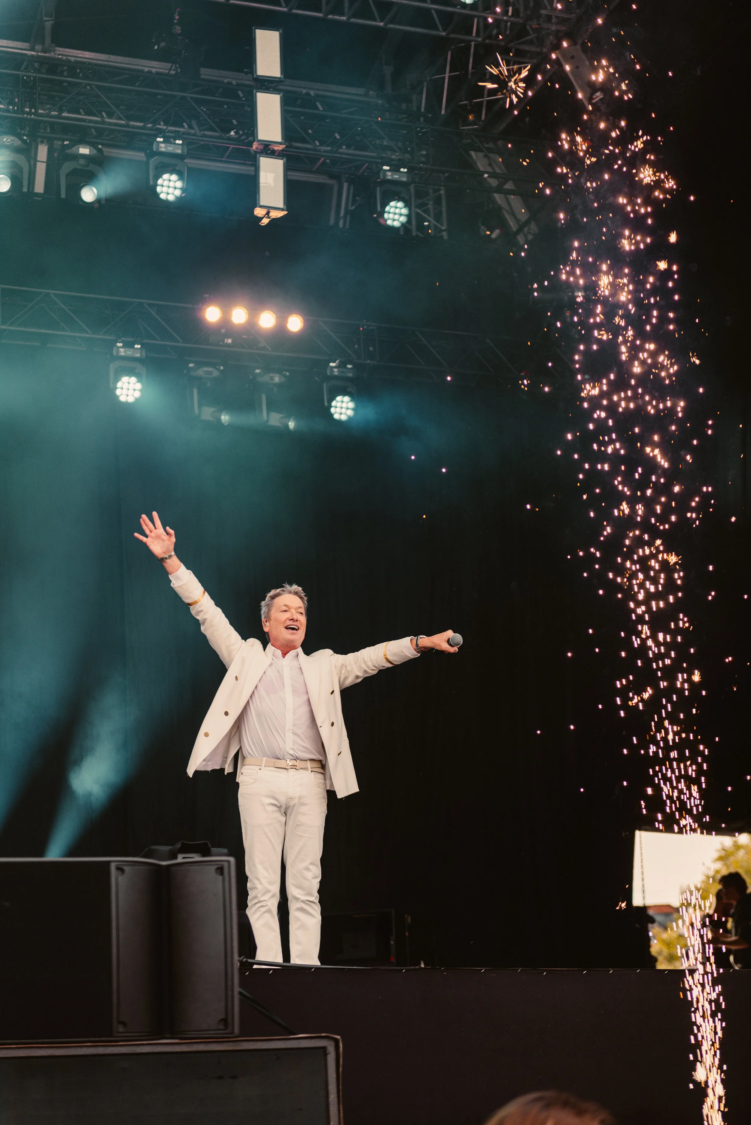 Performer in white suit on stage, holding a microphone, with arms raised, surrounded by stage lights and pyrotechnics.