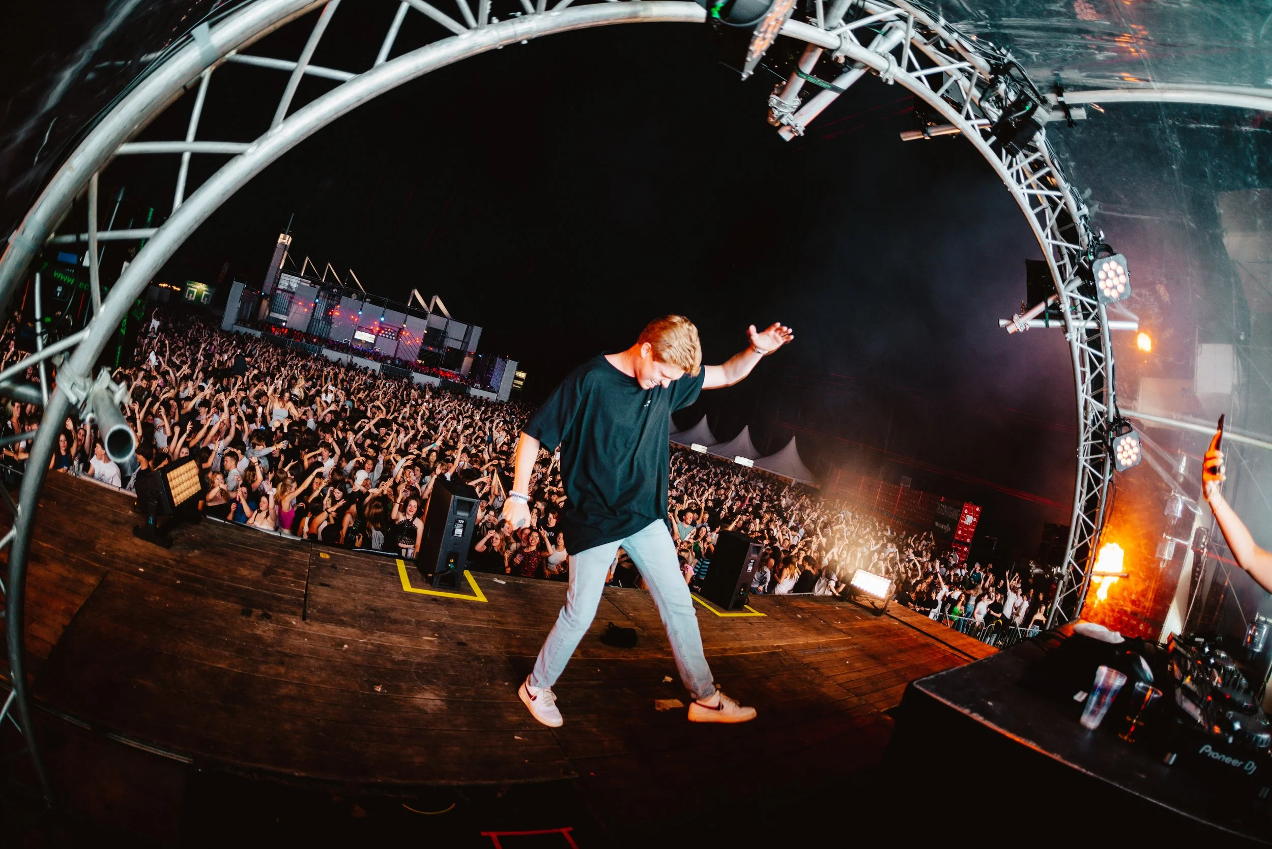 DJ performing on stage with large crowd at outdoor music festival, night scene