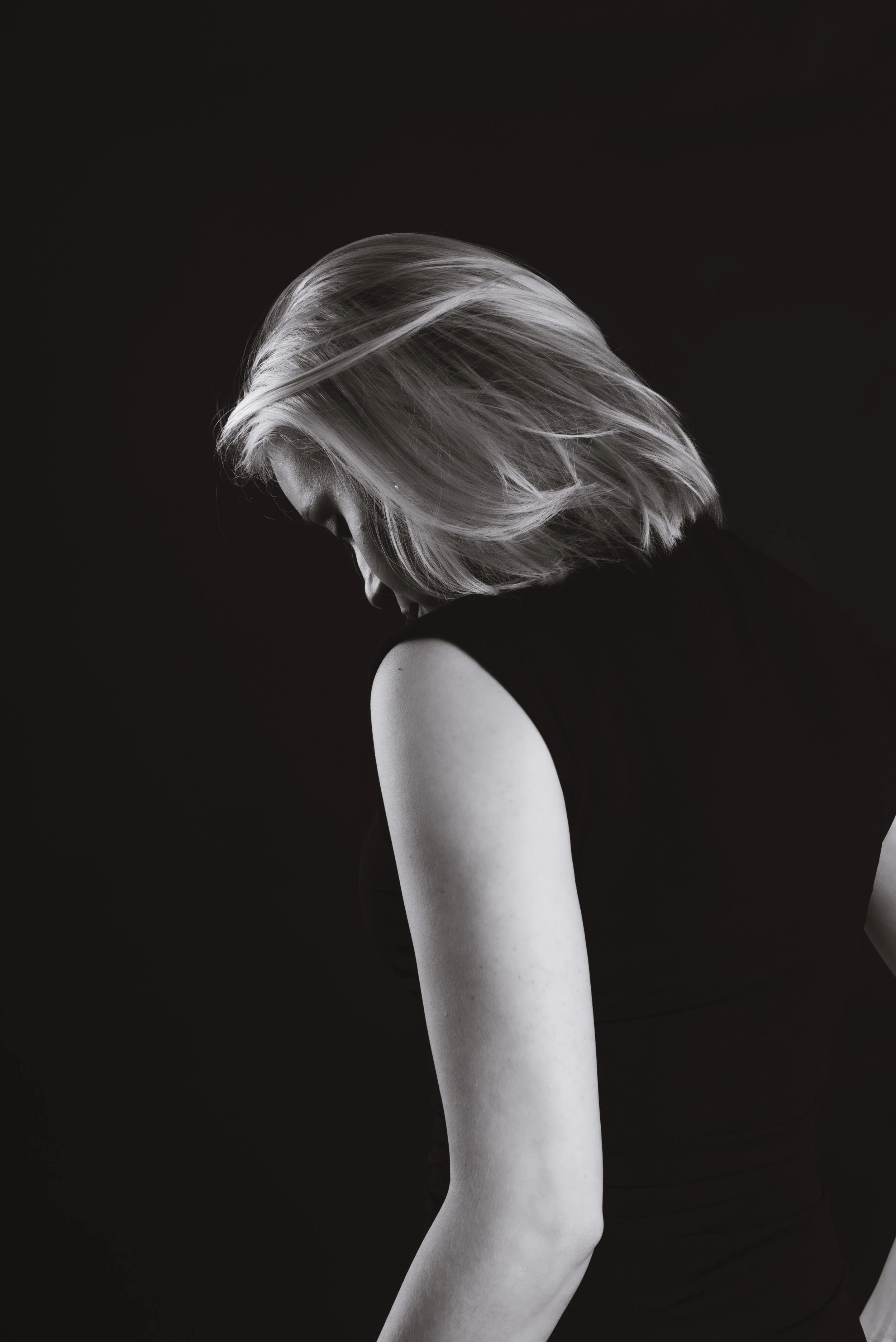 Black and white photo of a woman with shoulder-length hair looking down, wearing a sleeveless black top against a dark background.