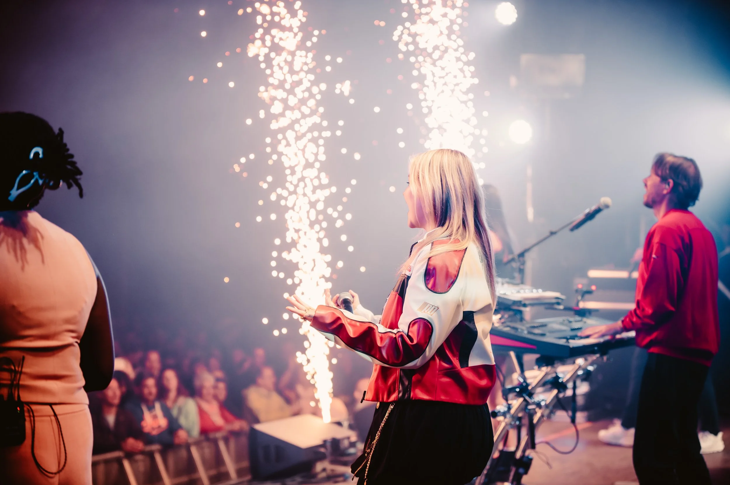 Musicians performing on stage with pyrotechnics and an audience in the background.