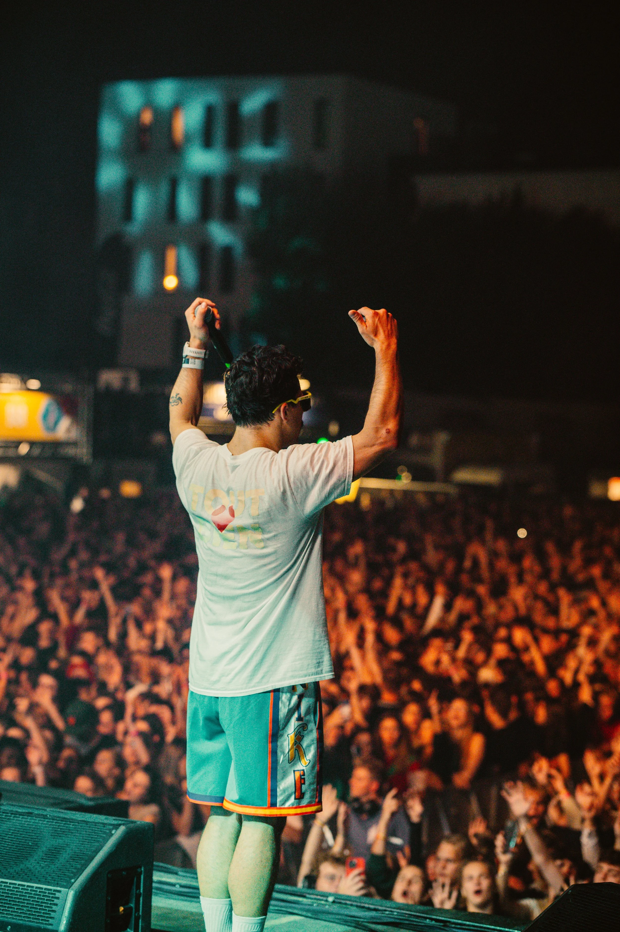 Performer on stage with a large crowd, wearing a white shirt and blue shorts, arms raised.