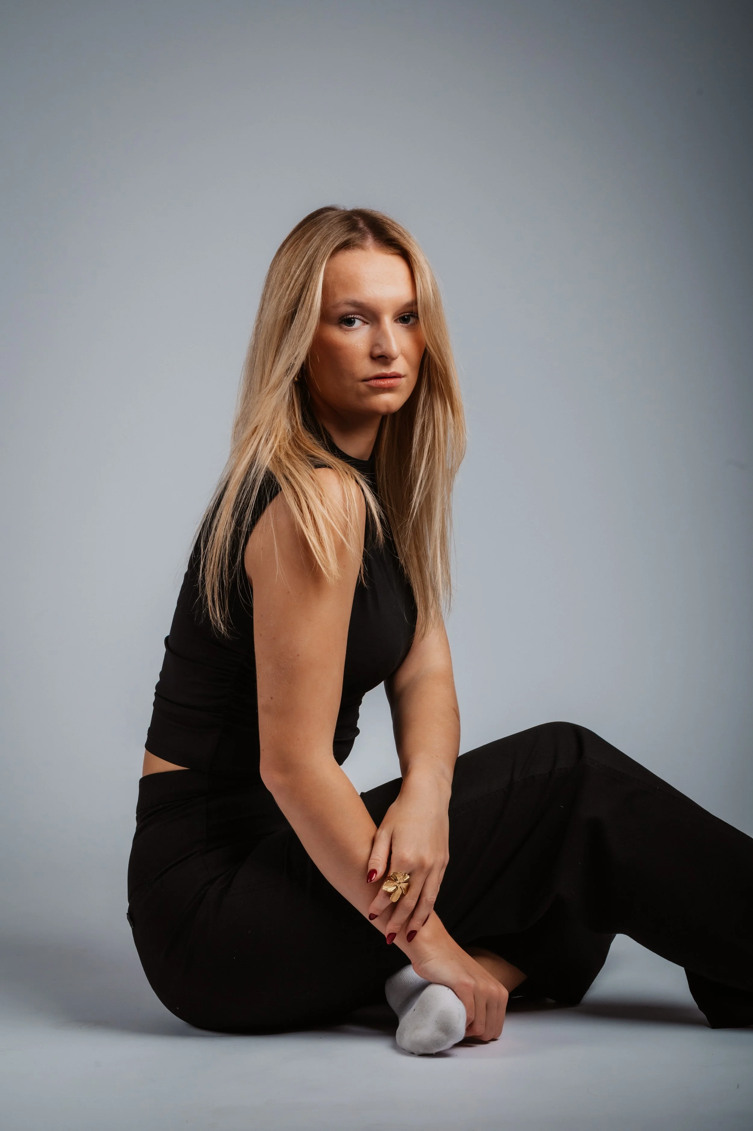 Woman with long blonde hair wearing a black sleeveless outfit, sitting on the floor against a neutral background.