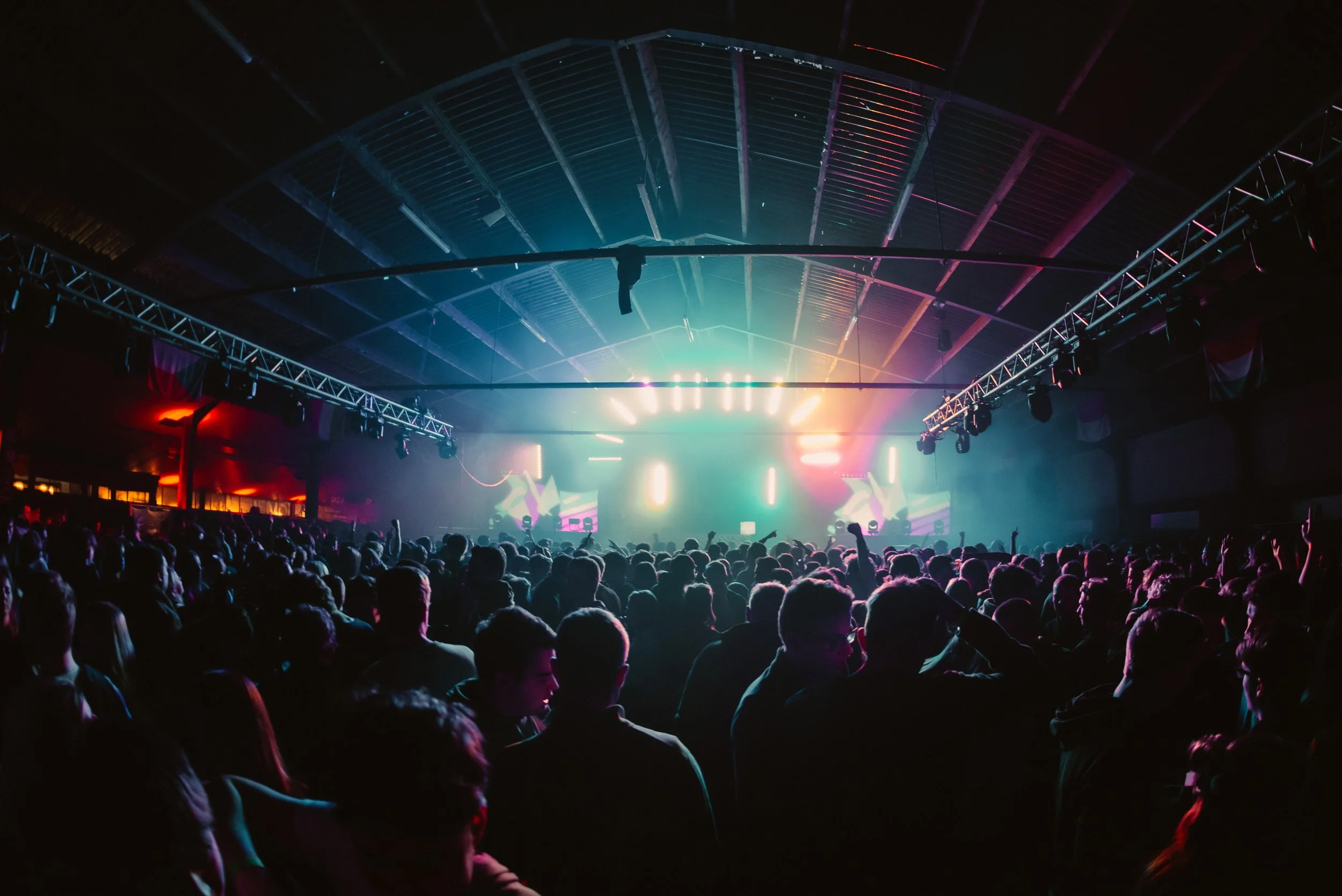 Large crowd enjoying a concert with colorful stage lights in a dark indoor venue.