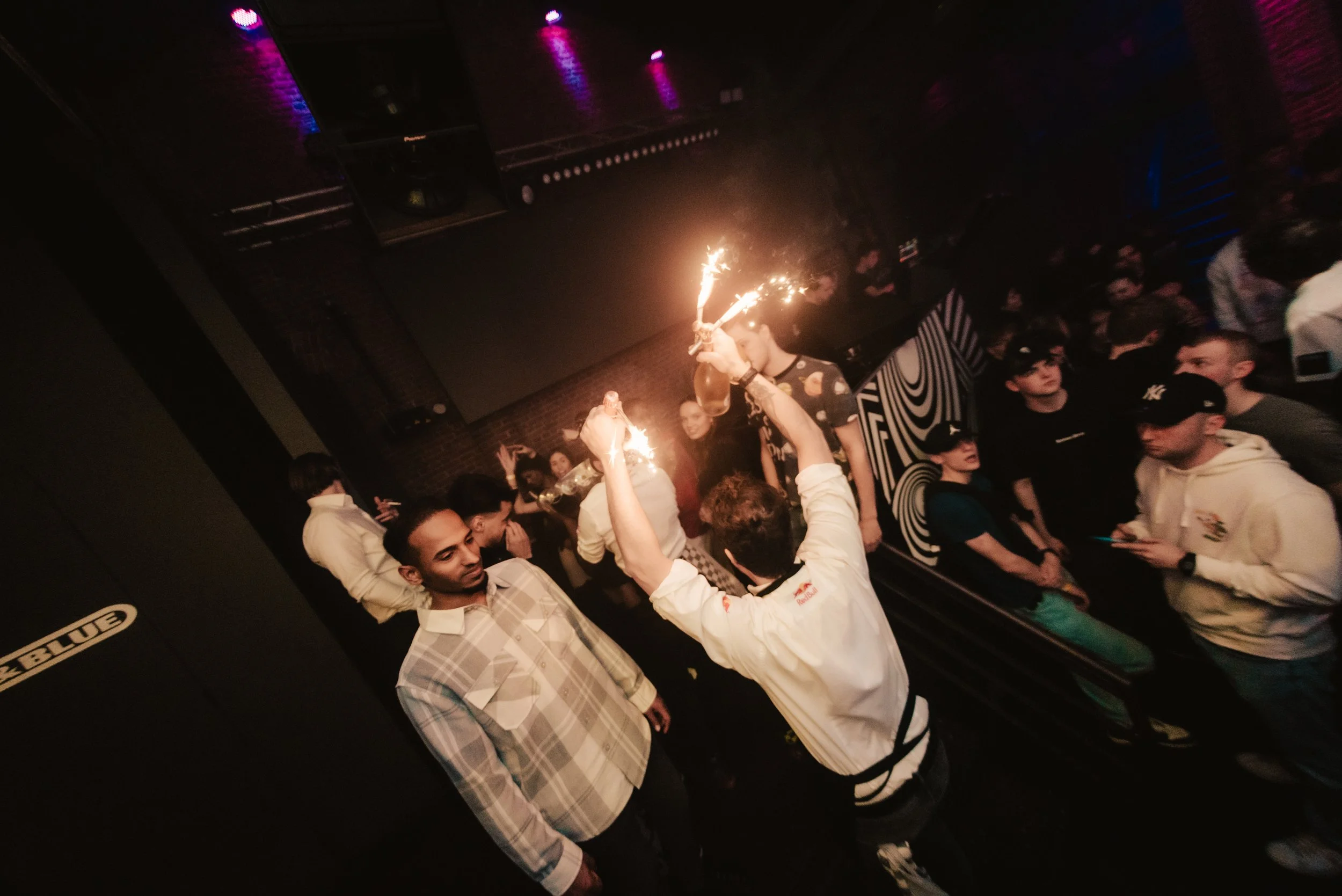 Crowded nightclub scene with a person holding sparklers, surrounded by people in casual clothing, under dim lighting with a backdrop of geometric wall patterns.