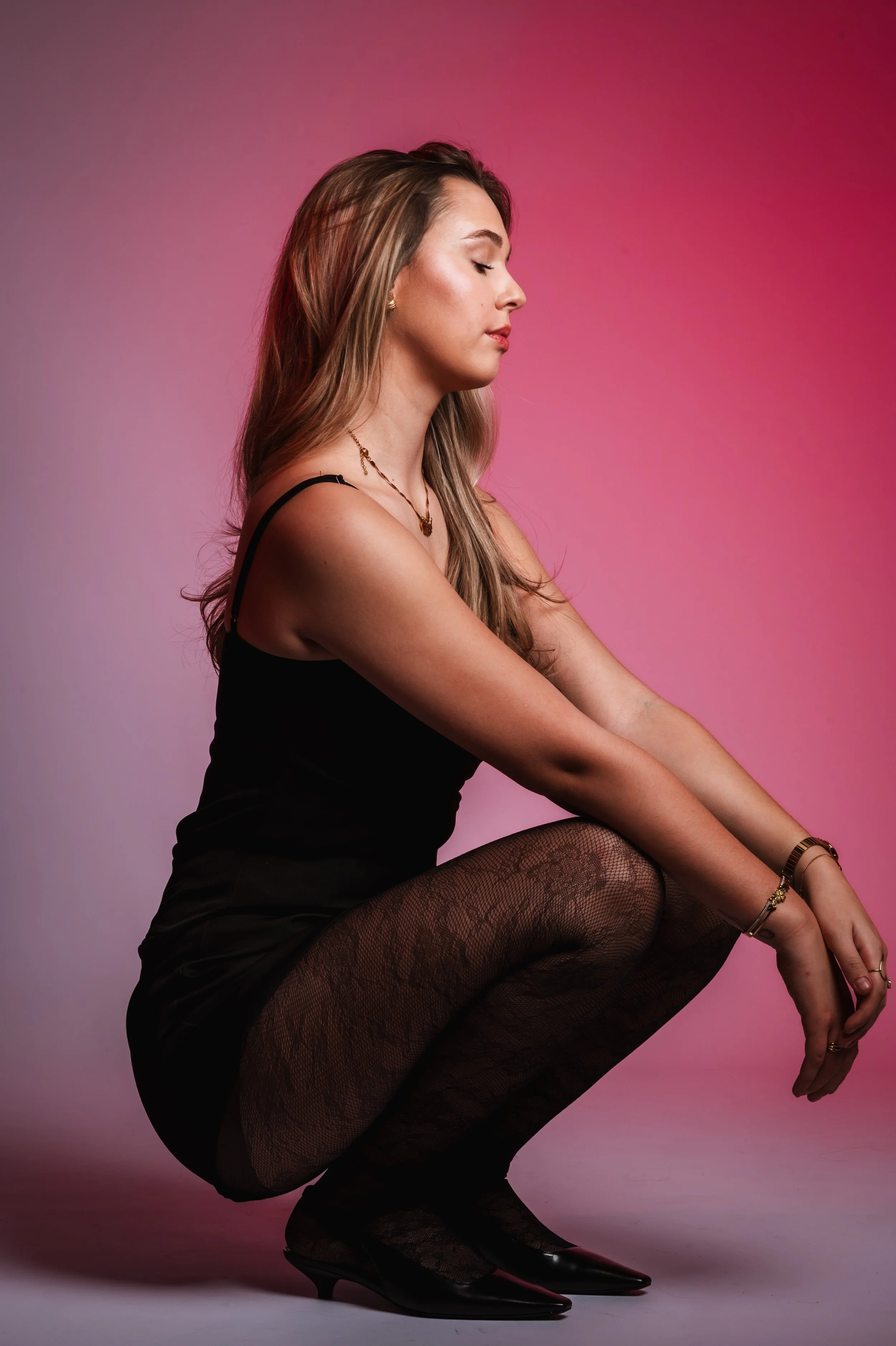 A young woman with long, wavy blonde hair is squatting, dressed in black, with eyes closed and a peaceful expression, against a pink gradient background.