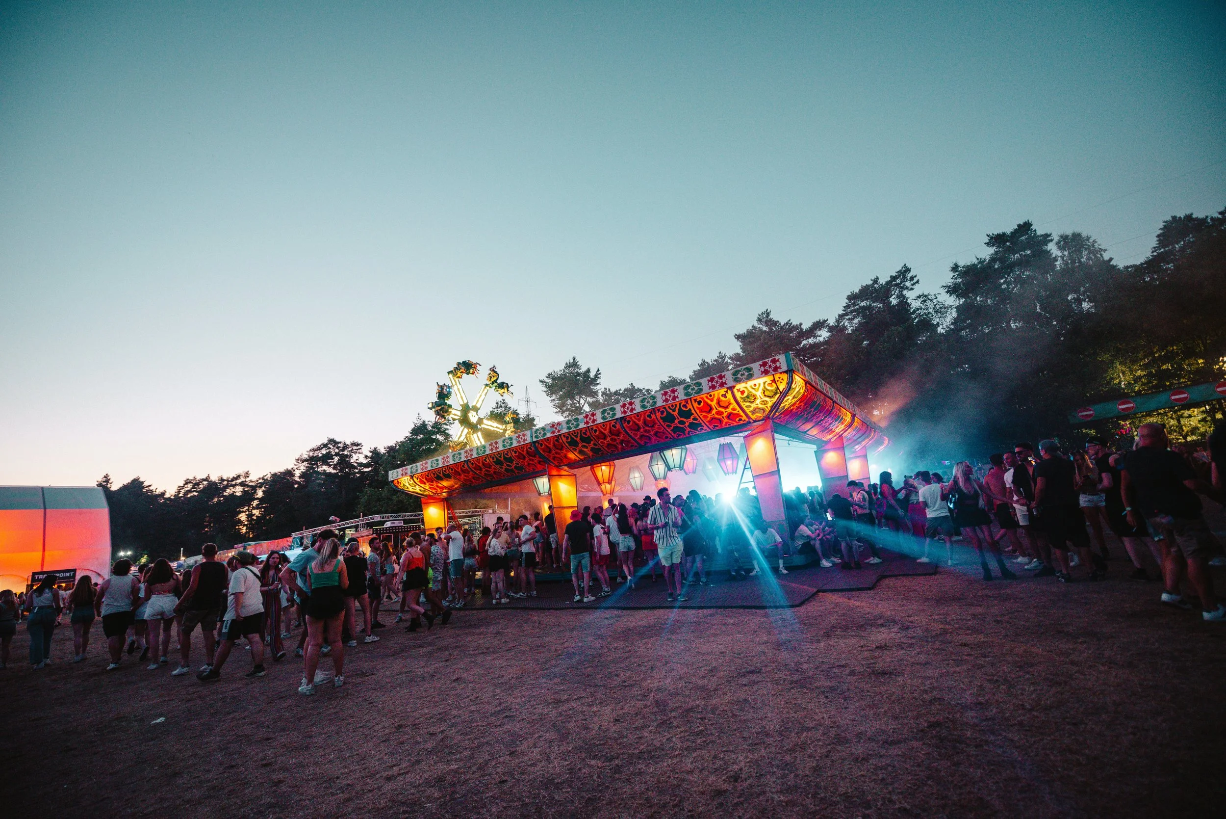 Outdoor festival with colorful lights, crowd of people, and a lit-up structure at sunset.