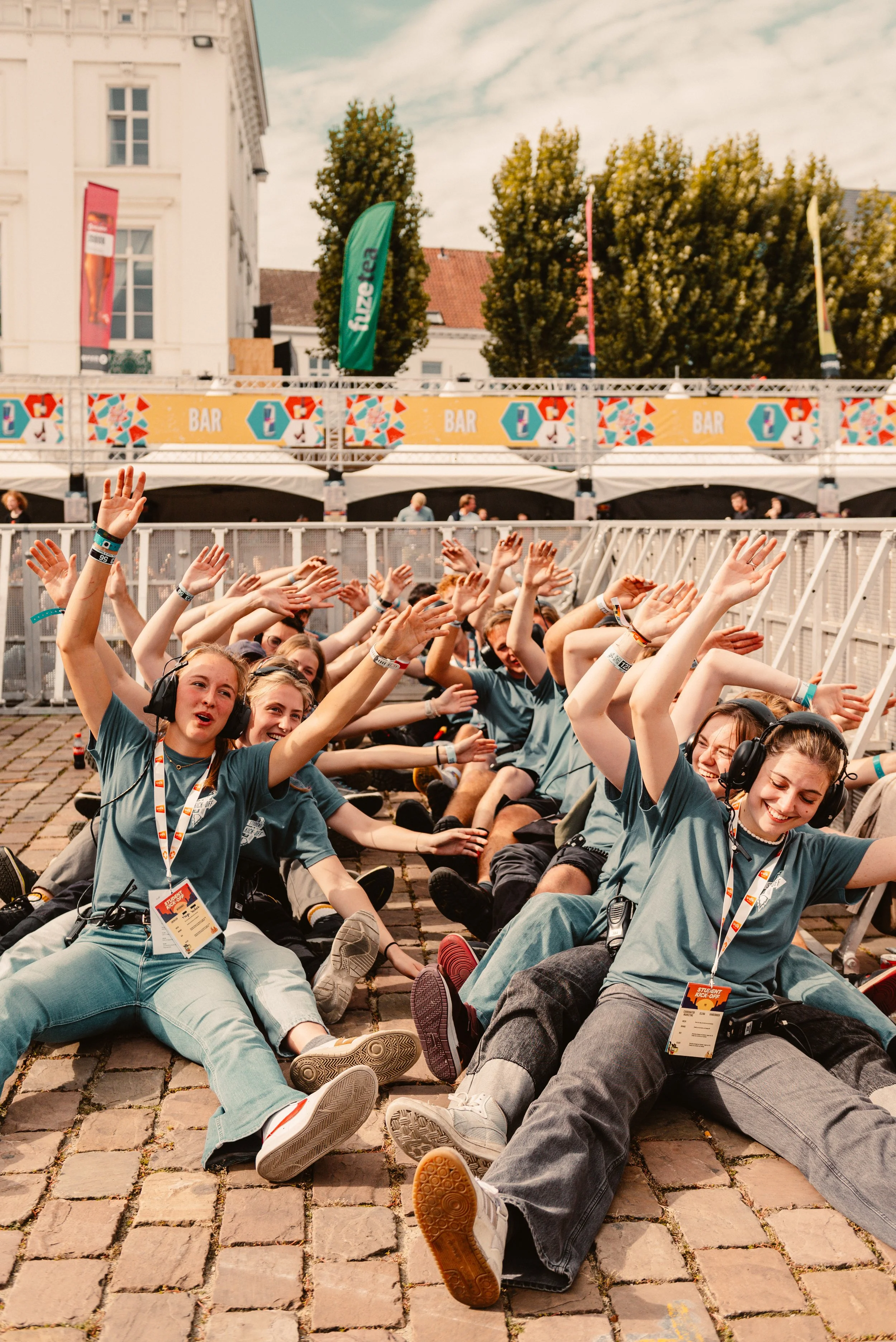 Group of people wearing blue shirts sitting on the ground, raising their arms, with event badges around their necks, in front of a bar area.