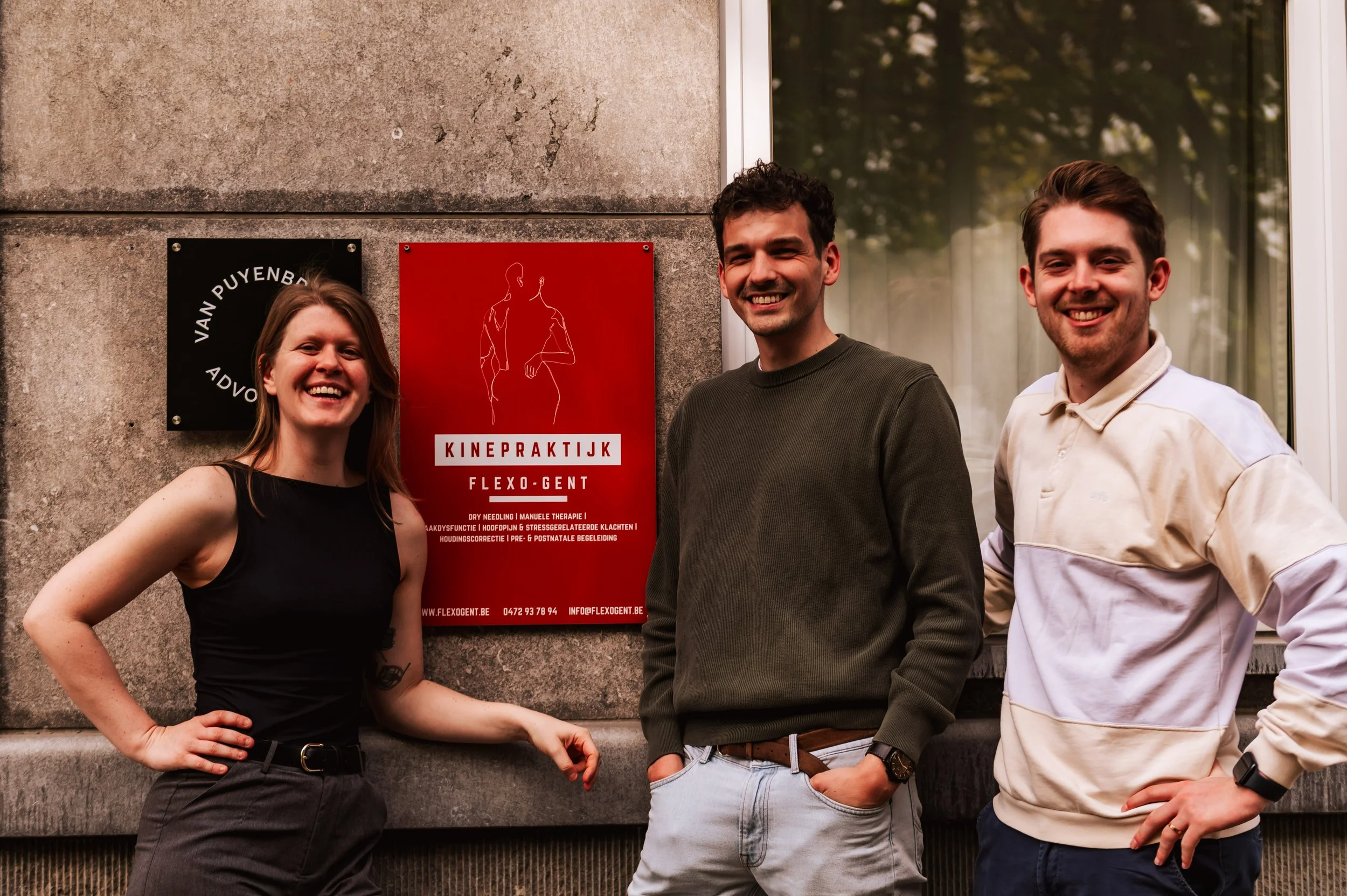 Three people smiling outside near a red sign labeled 'KINEPRAKTIJK FLEXO:GENT'.