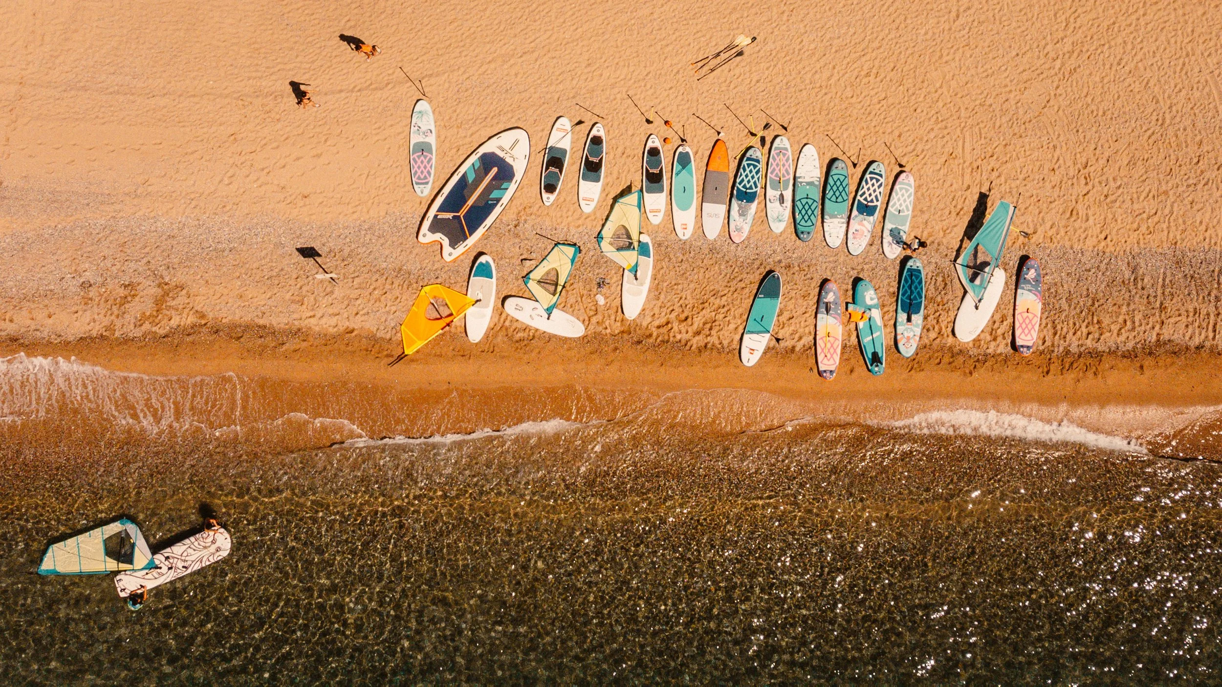 Aerial view of a beach with paddleboards lined up on the sand, a few with sails, and waves washing up on shore.