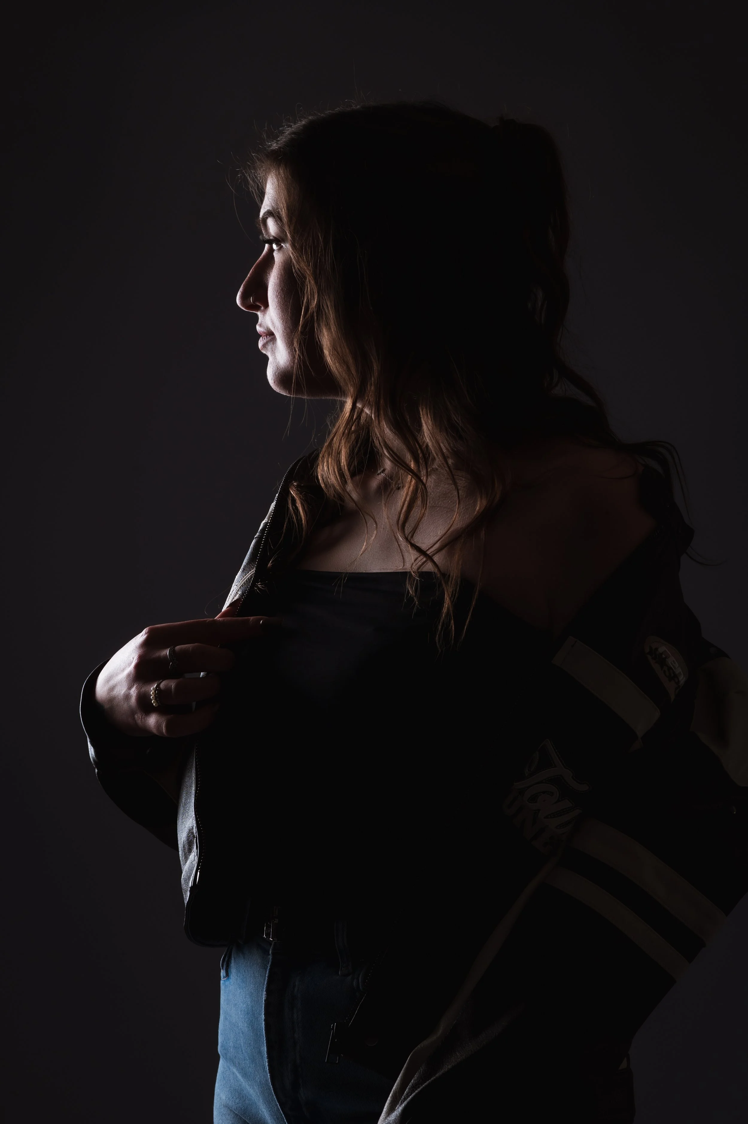 Side profile of a woman with wavy hair wearing a dark jacket and blue jeans, holding her collar, against a dark background.