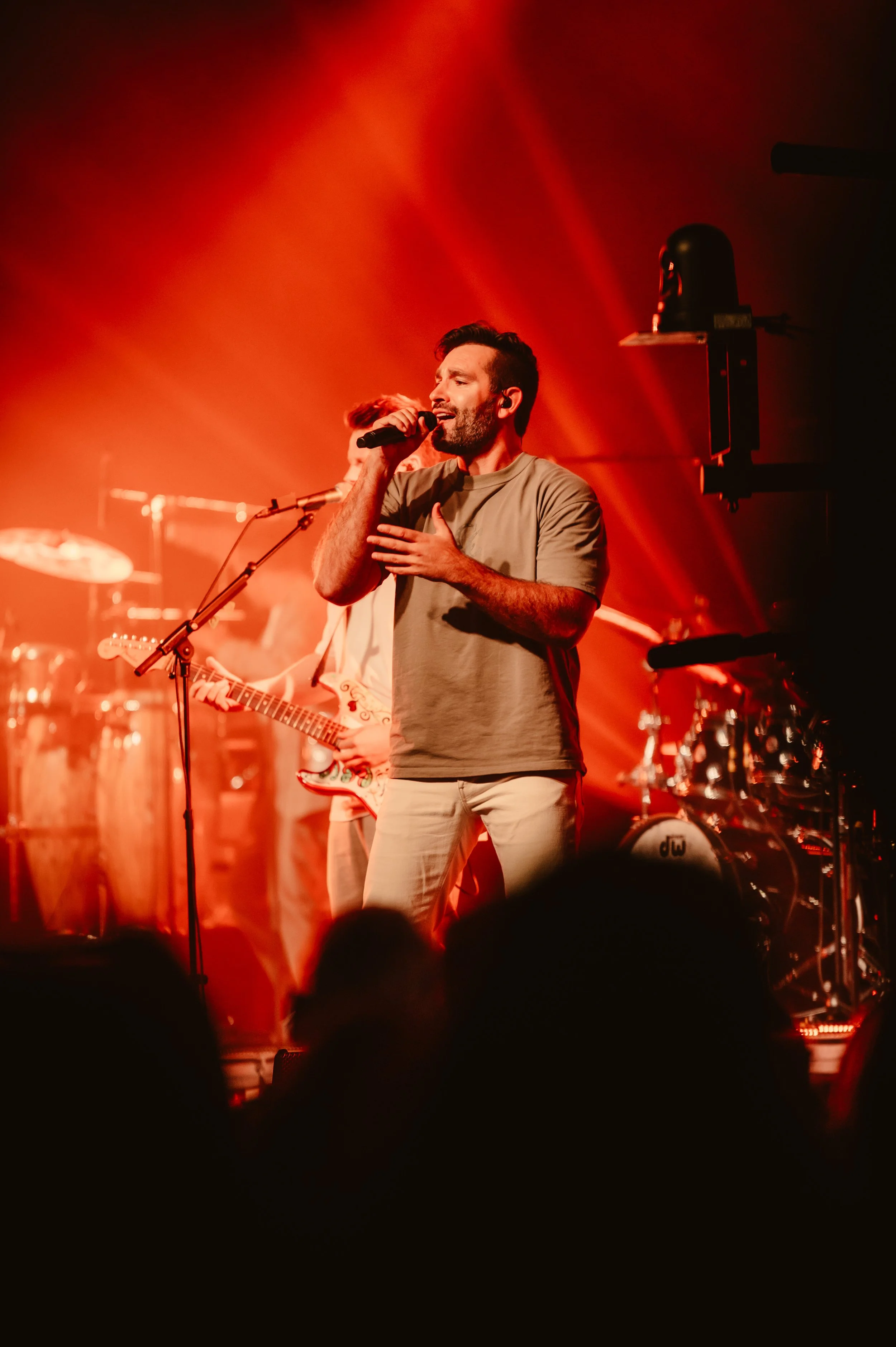 Singer performing on stage with microphone, surrounded by musical instruments and red stage lighting.