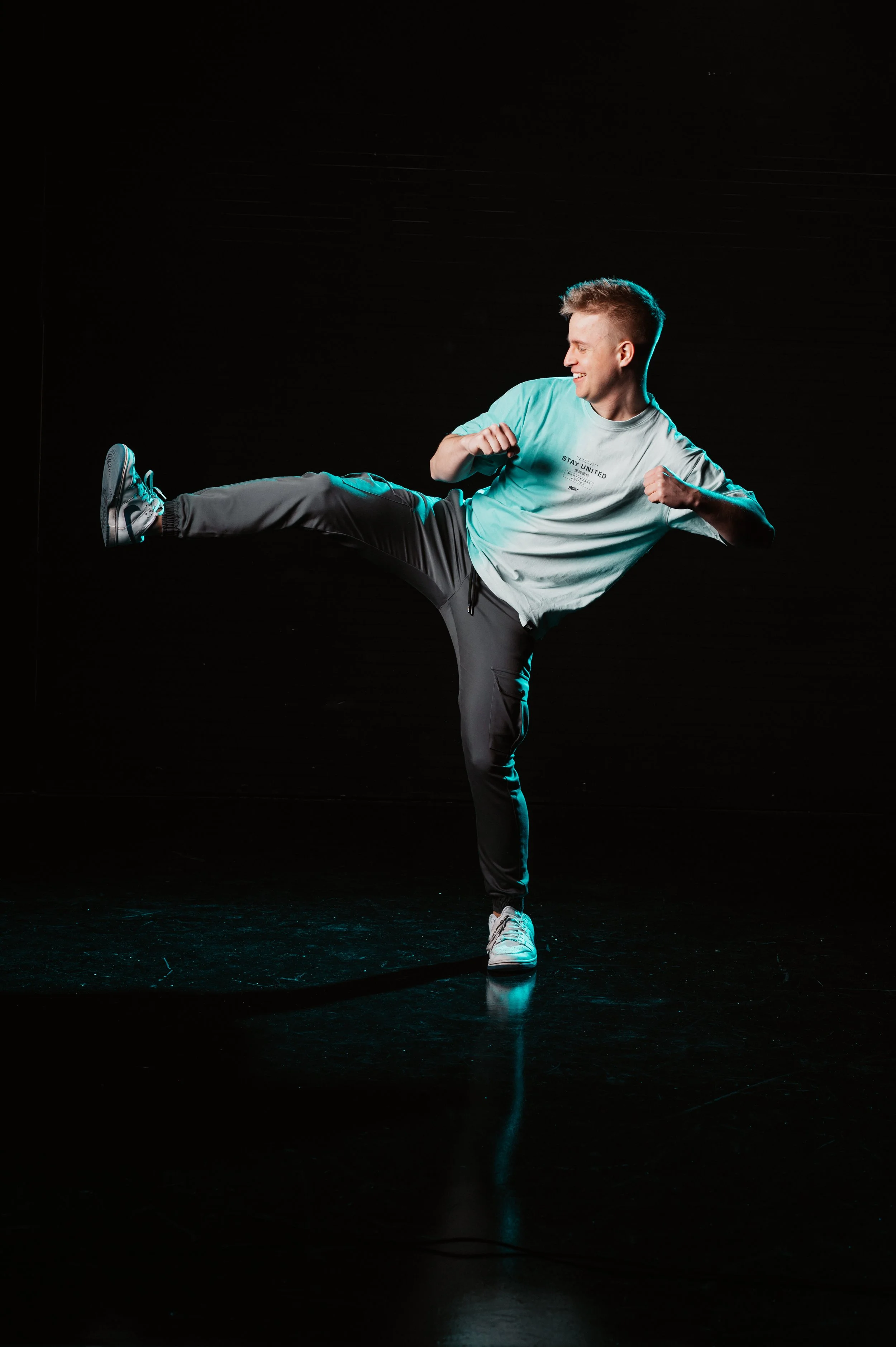 A person performing a high kick in a martial arts pose on a dark background, wearing a light t-shirt and dark pants.