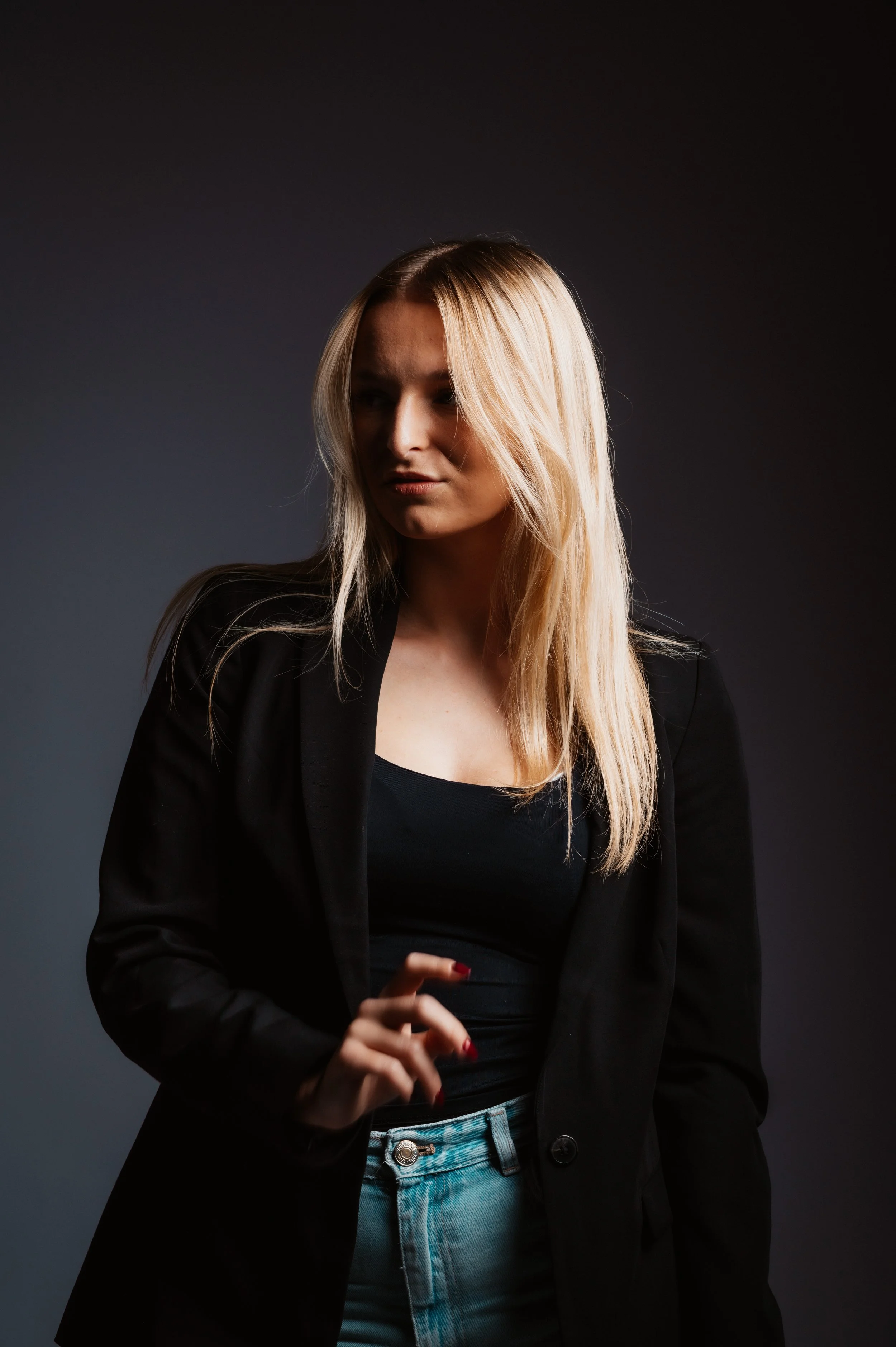 Blonde woman in black blazer and jeans posing against a dark background.