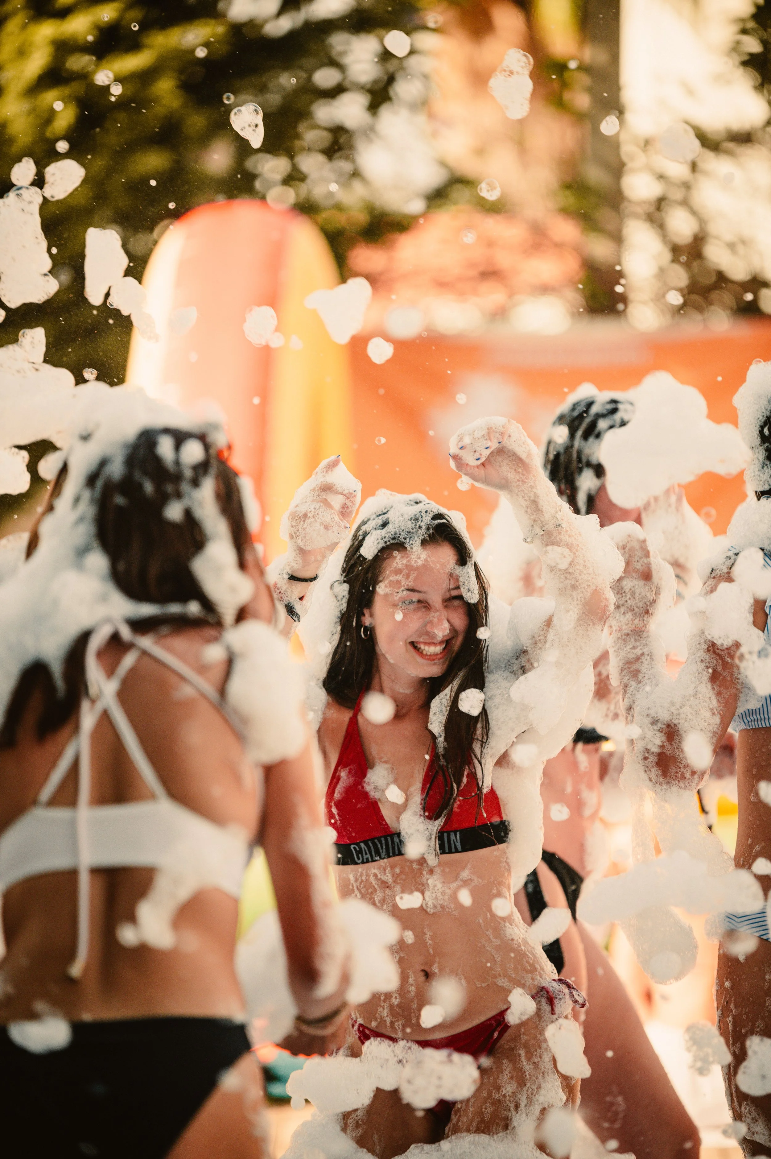 Group of people enjoying a foam party outdoors, wearing swimsuits and covered in bubbles, with trees in the background.