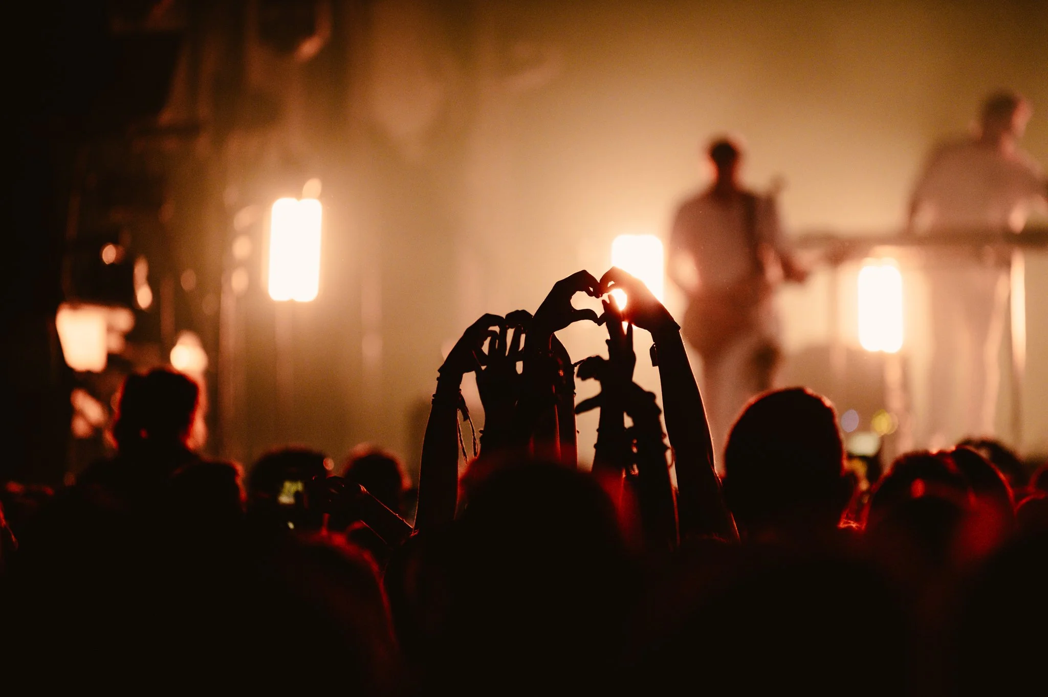 Concert crowd with hands forming heart shapes, artists performing on stage, bright stage lights in background.