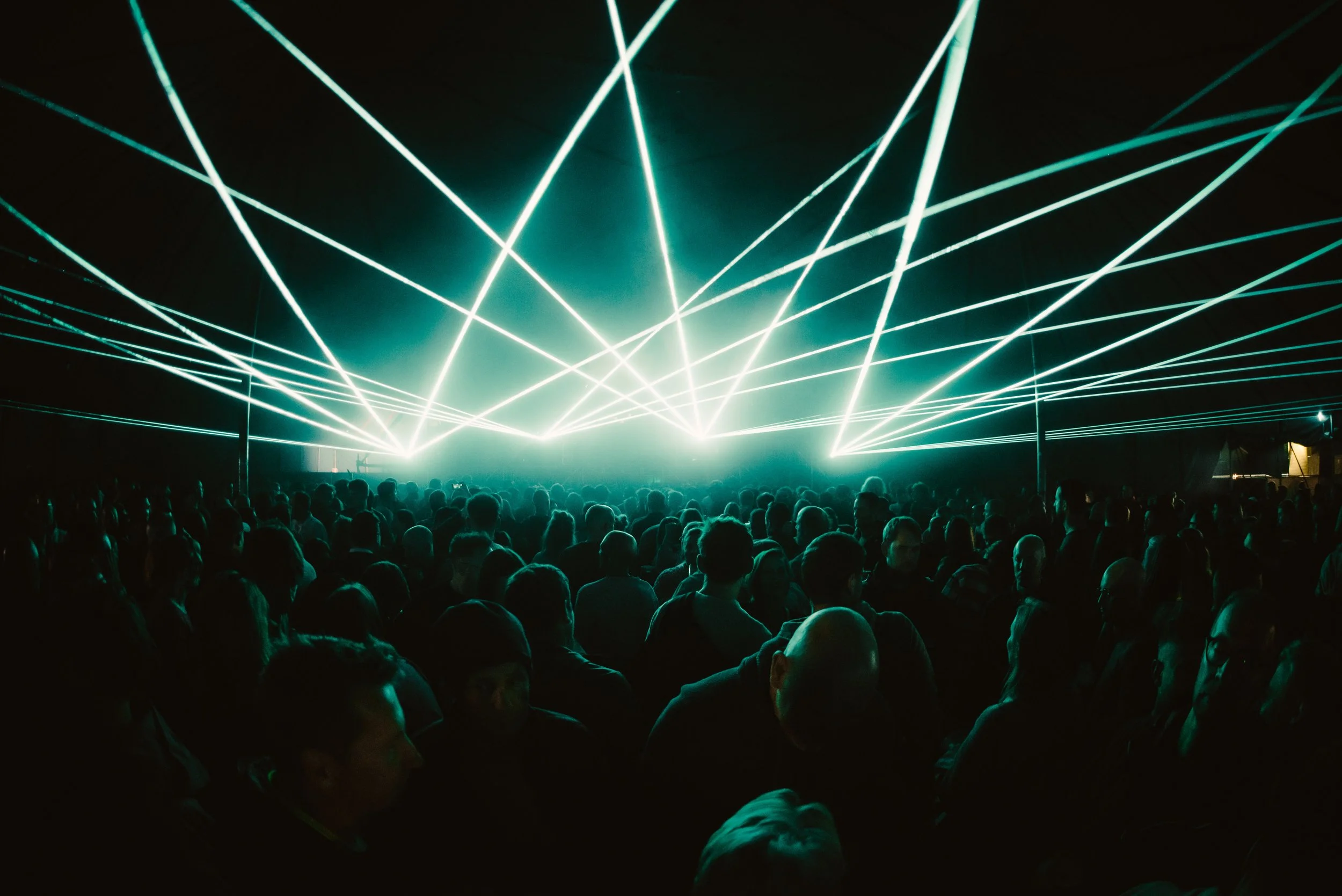 Crowd of people at a concert with blue laser lights overhead, creating a vibrant atmosphere.