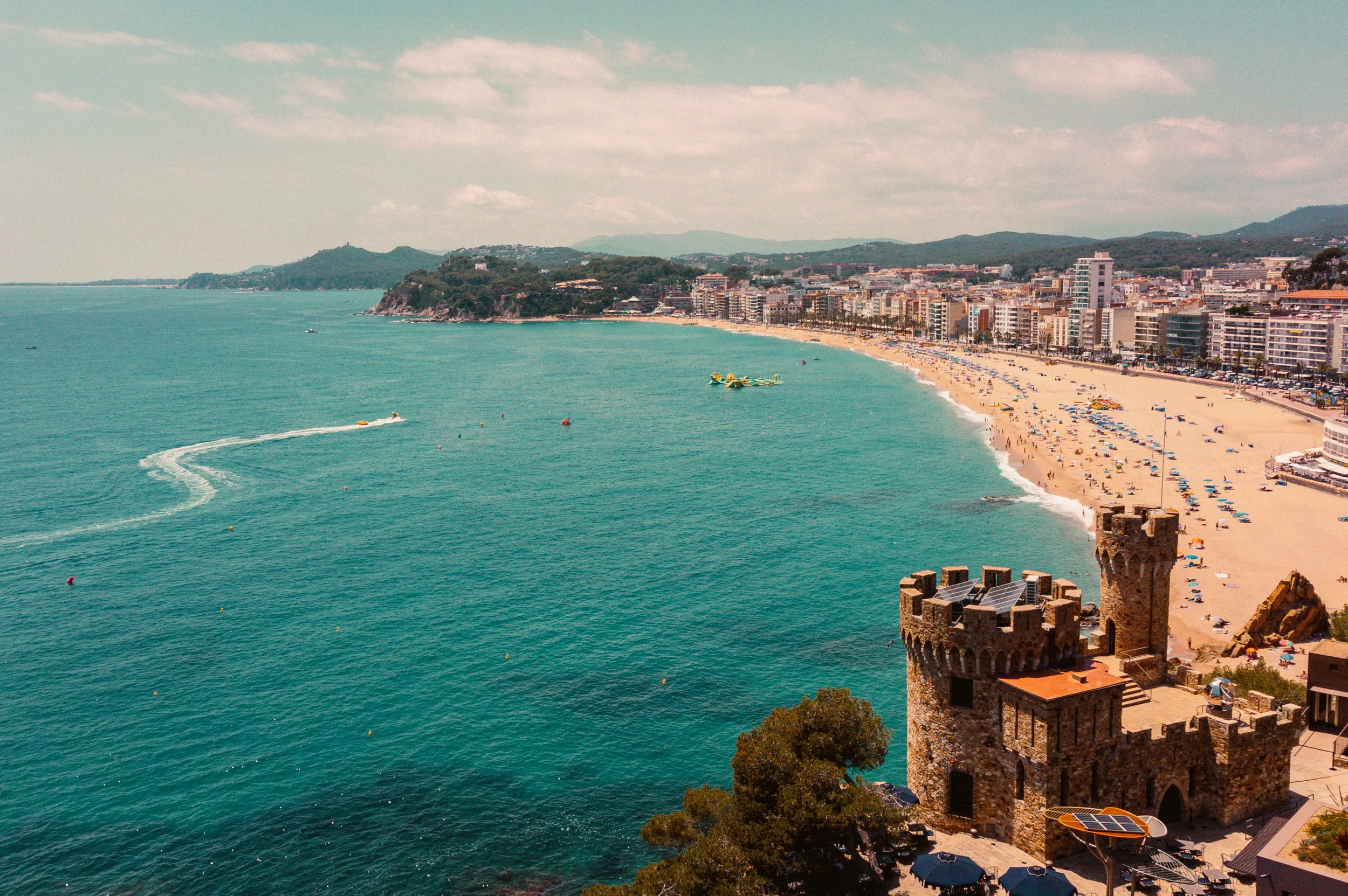 Aerial view of a beach with turquoise water, a castle-like tower in the foreground, and a city with high-rise buildings along the coastline.