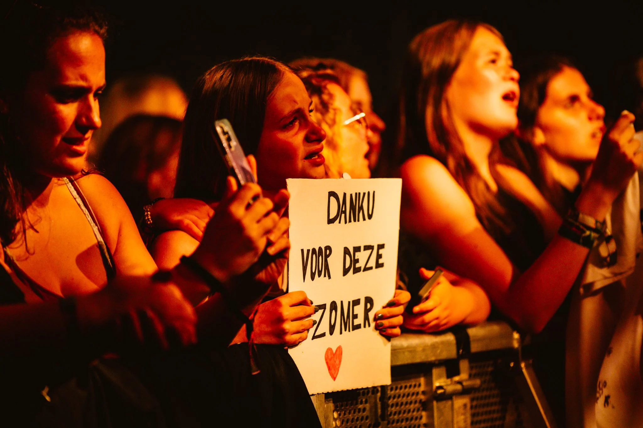 Crowd of excited concertgoers holding a sign reading "Danku voor deze zomer" with a heart, capturing the lively atmosphere of a music event.