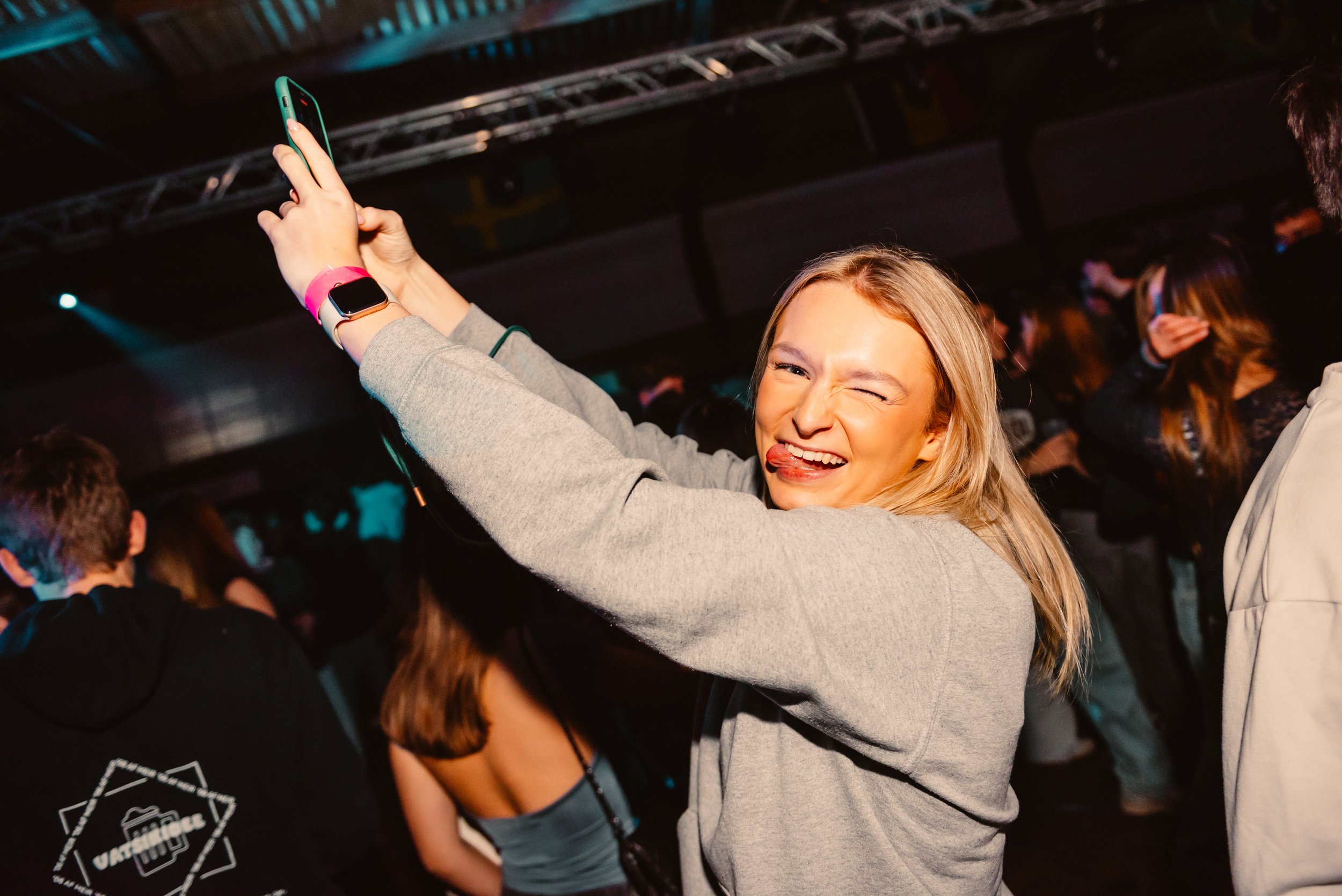 A woman smiling and taking a selfie in a crowded party or concert setting, wearing a gray sweatshirt, with people dancing in the background.