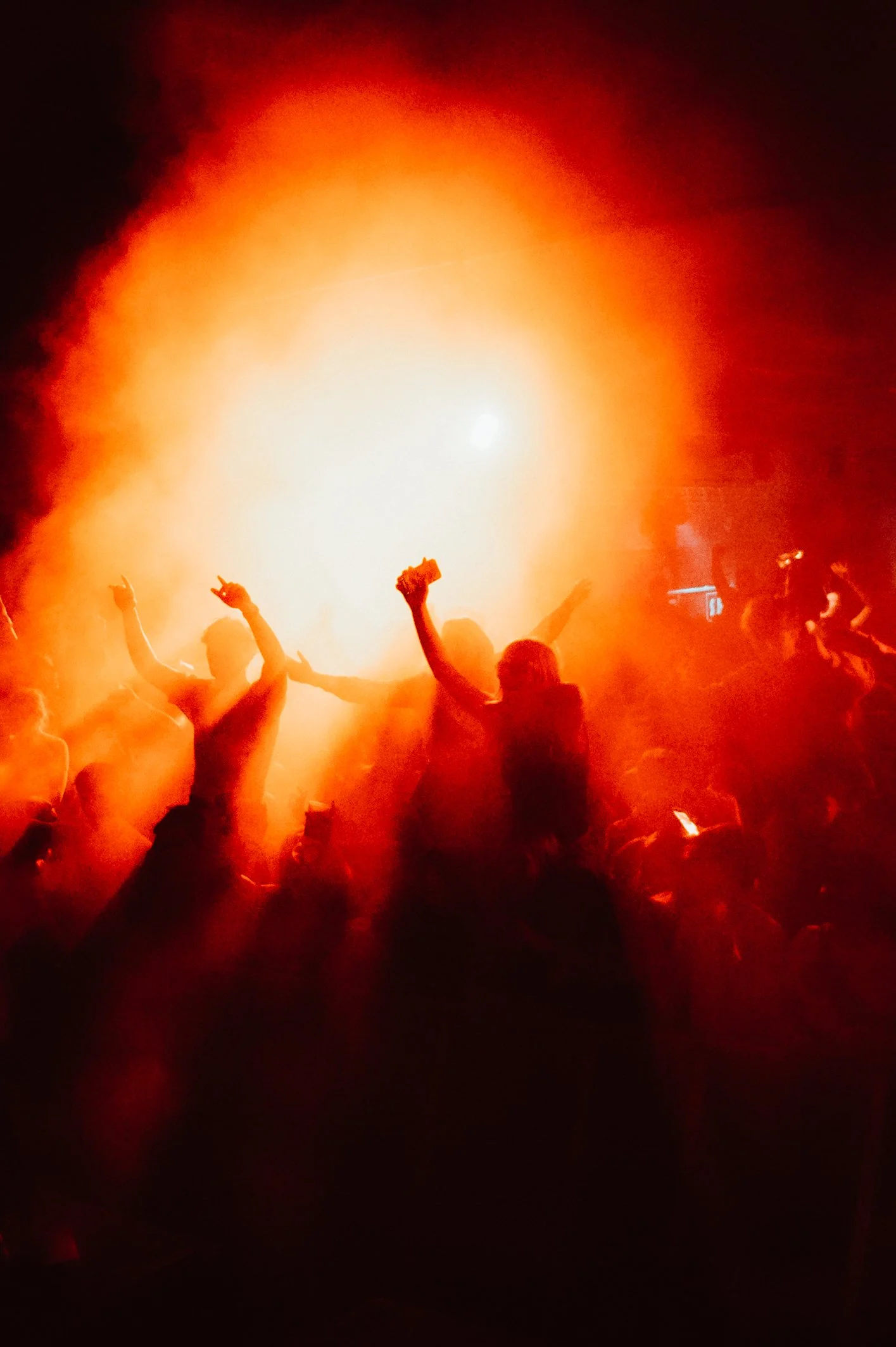 Silhouettes of people dancing with hands raised amidst red smoke and bright light at a concert or party.
