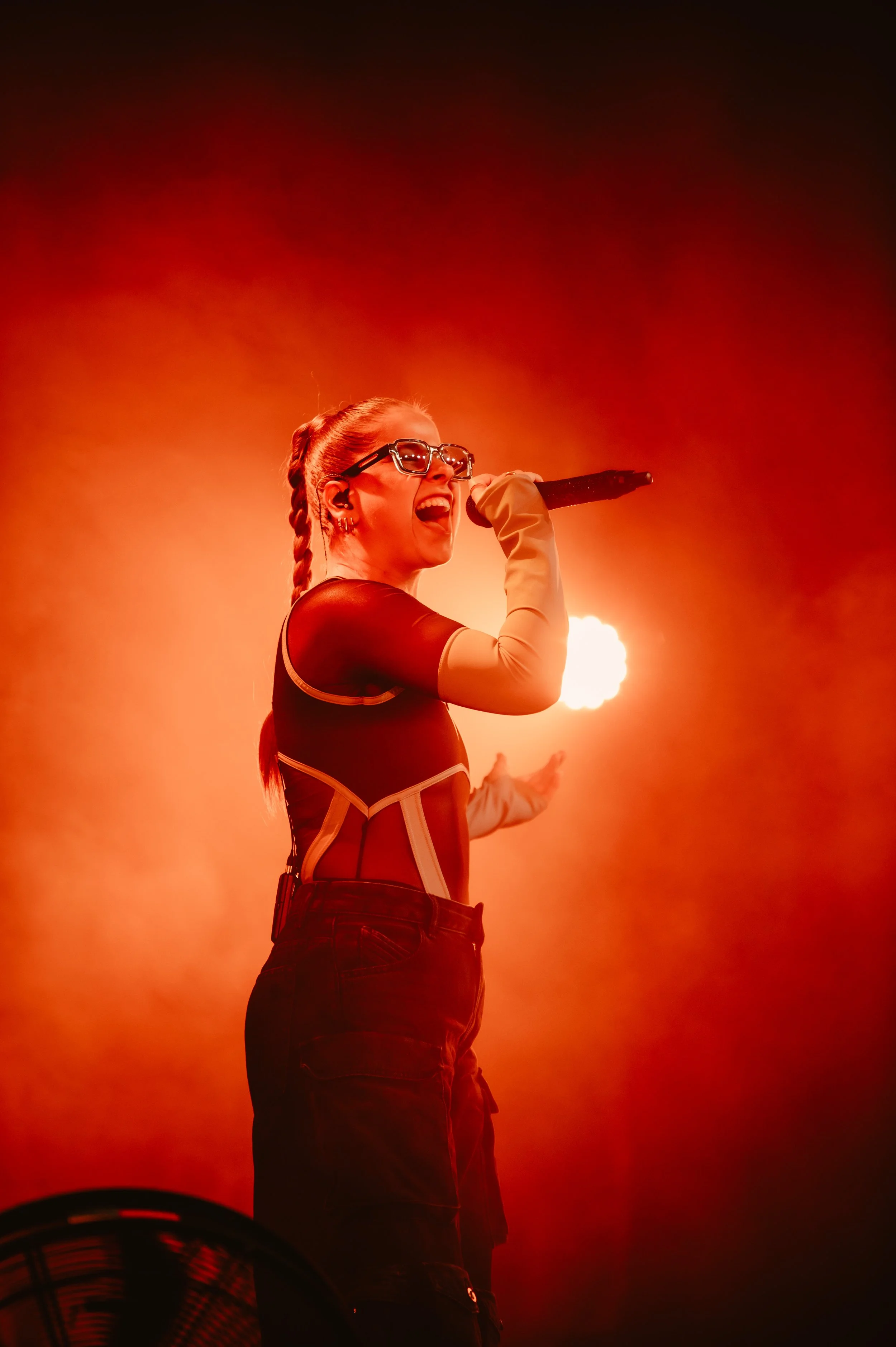 Performer singing energetically on stage with a microphone, wearing glasses and a stylish outfit, against a vibrant red-orange background.