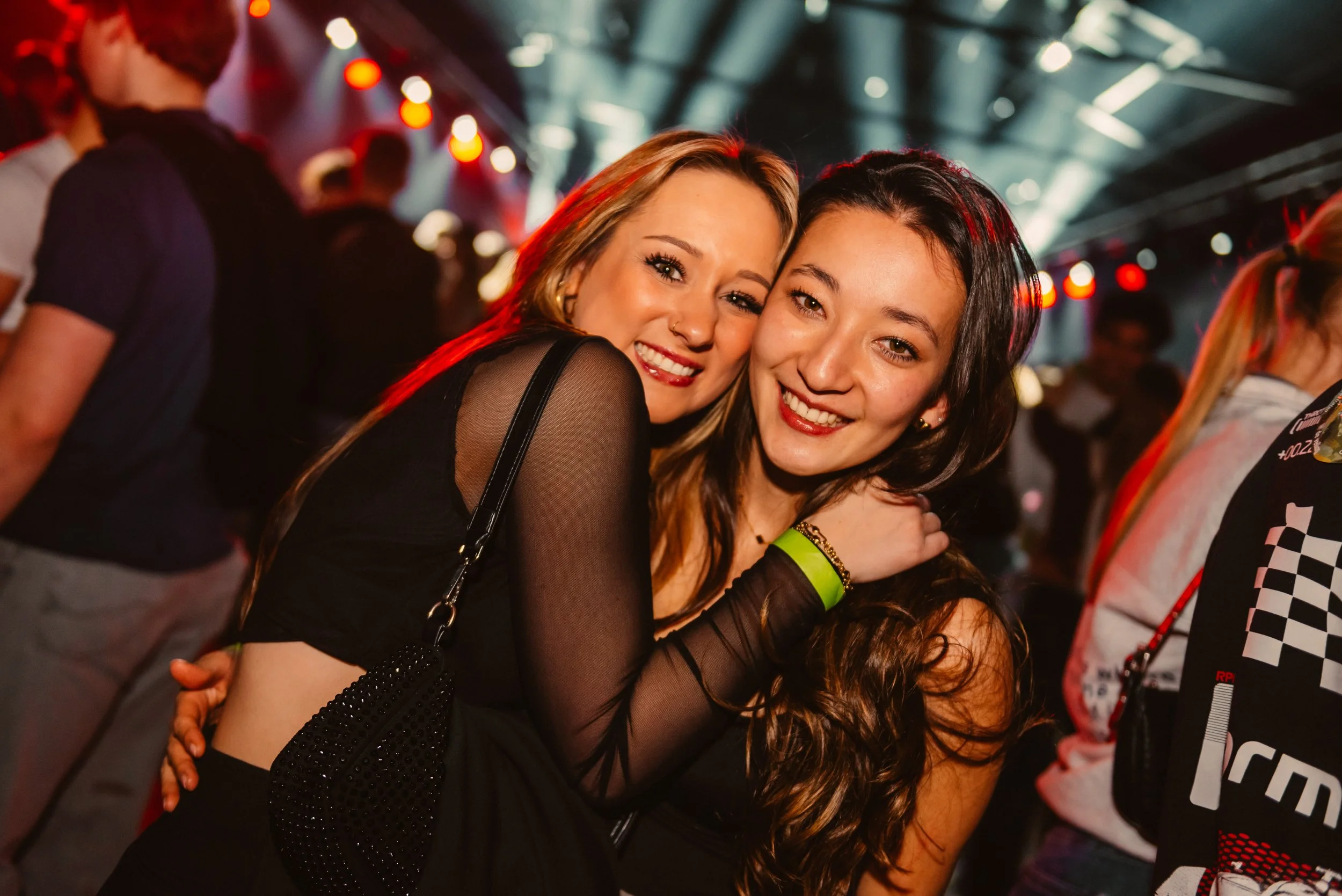 Two women smiling and embracing at a lively party with colorful lights and people in the background.