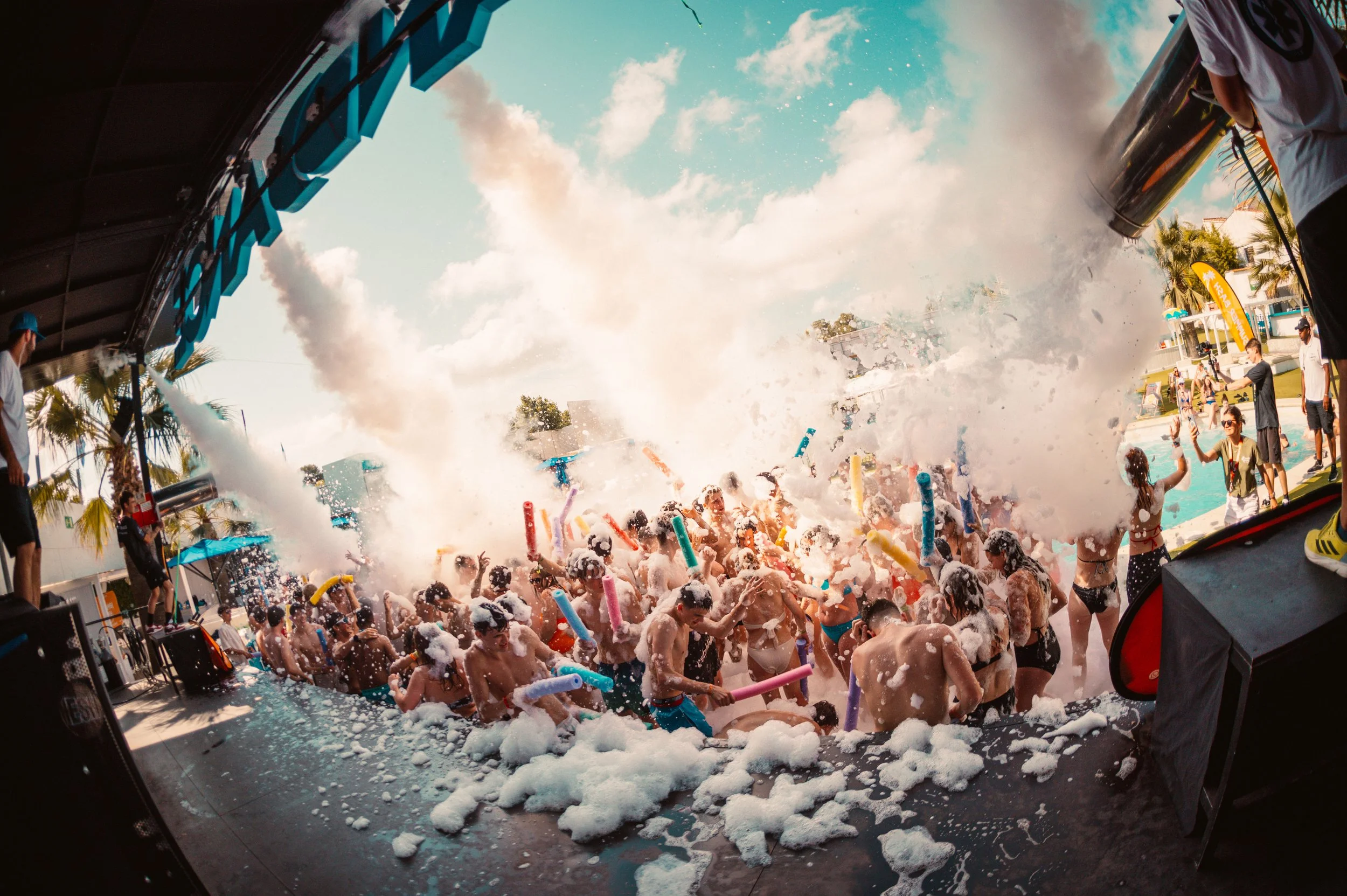 People enjoying a foam party outdoors, with foam machines spraying bubbles and individuals holding inflatable toys.