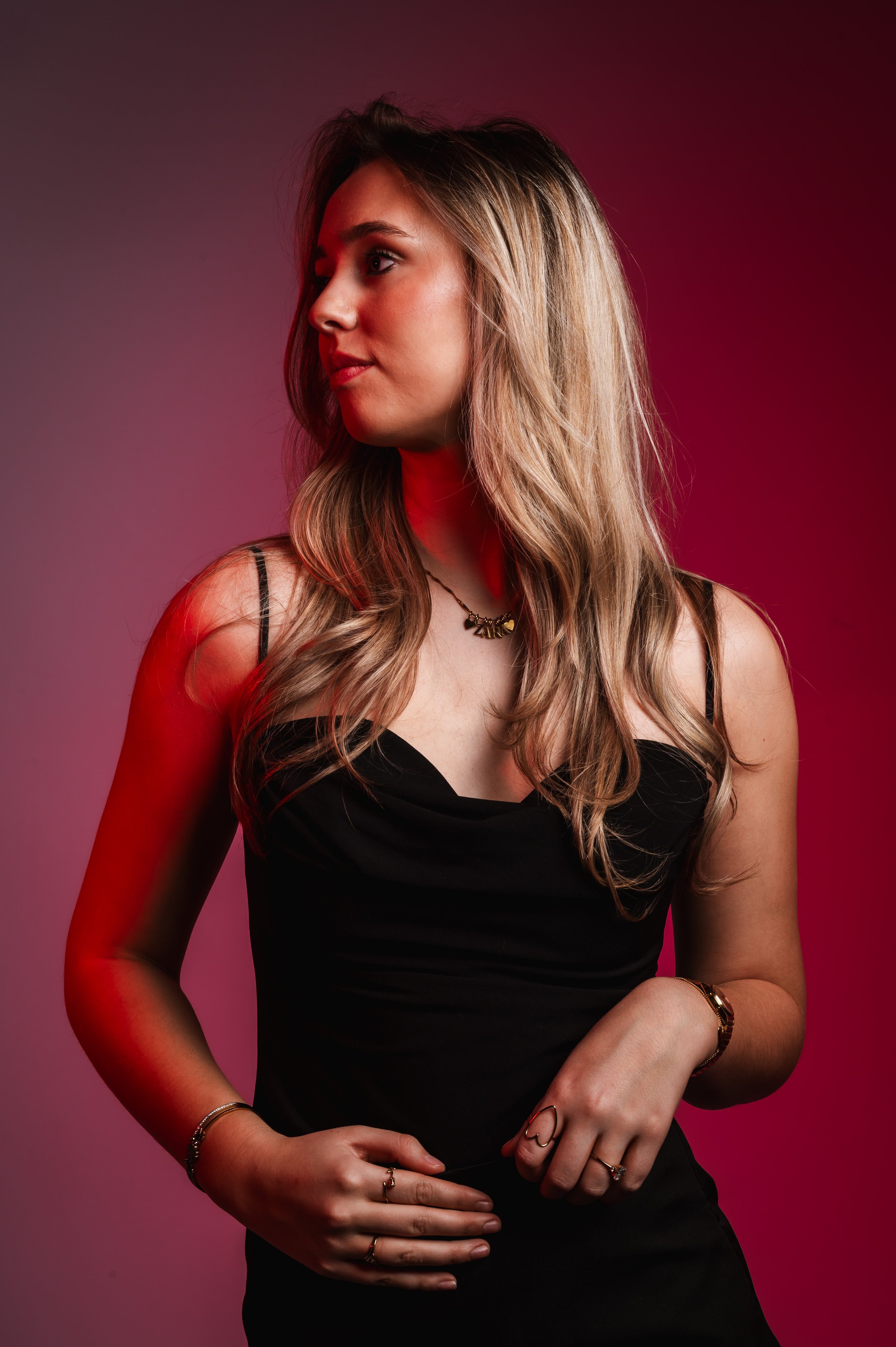 A young woman with long wavy blonde hair, wearing a black spaghetti strap top, jewelry, and standing against a pink and black gradient background.