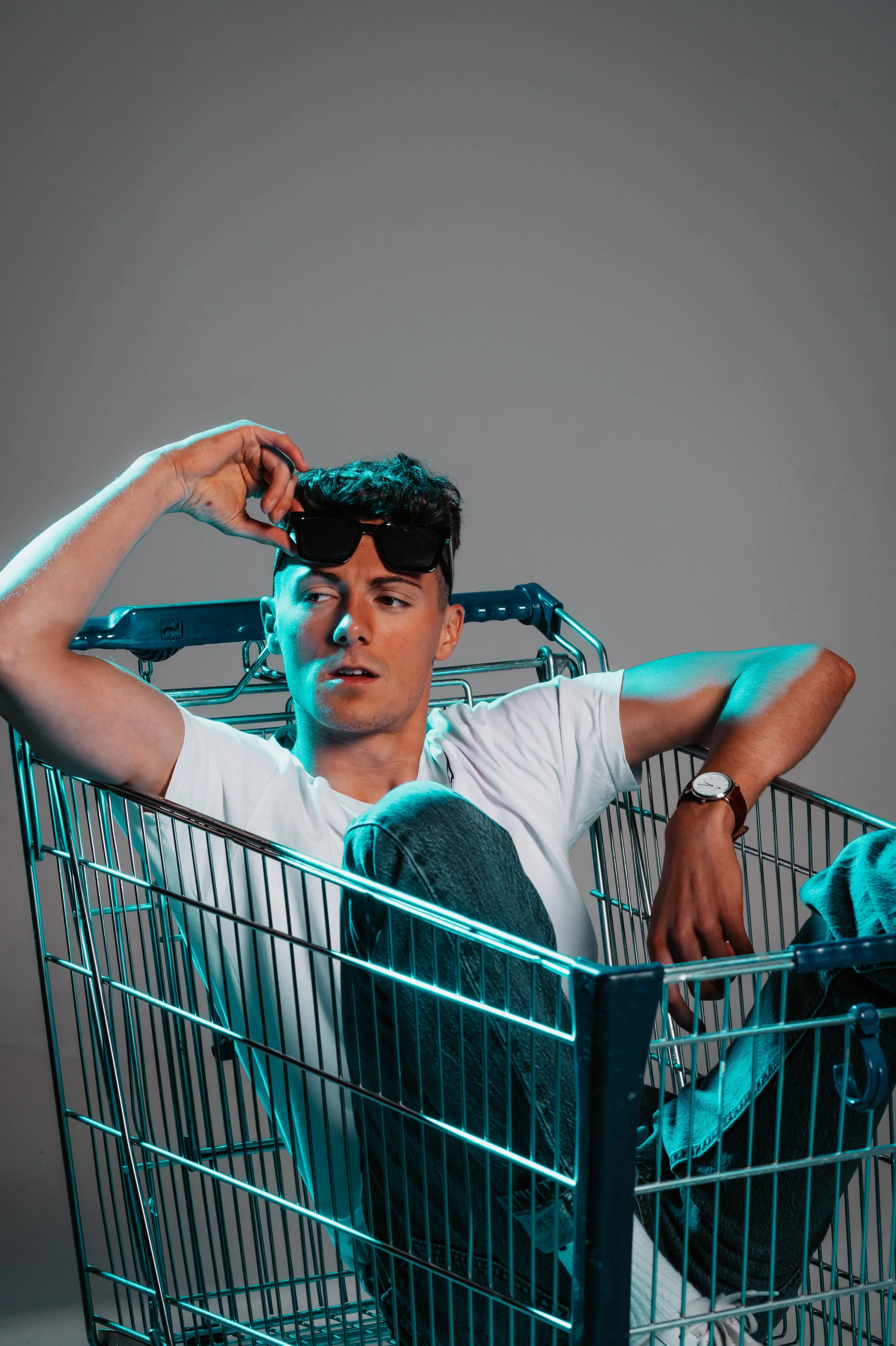 Person sitting in a shopping cart, wearing a white t-shirt, jeans, and sunglasses, with a wristwatch, under blue lighting.