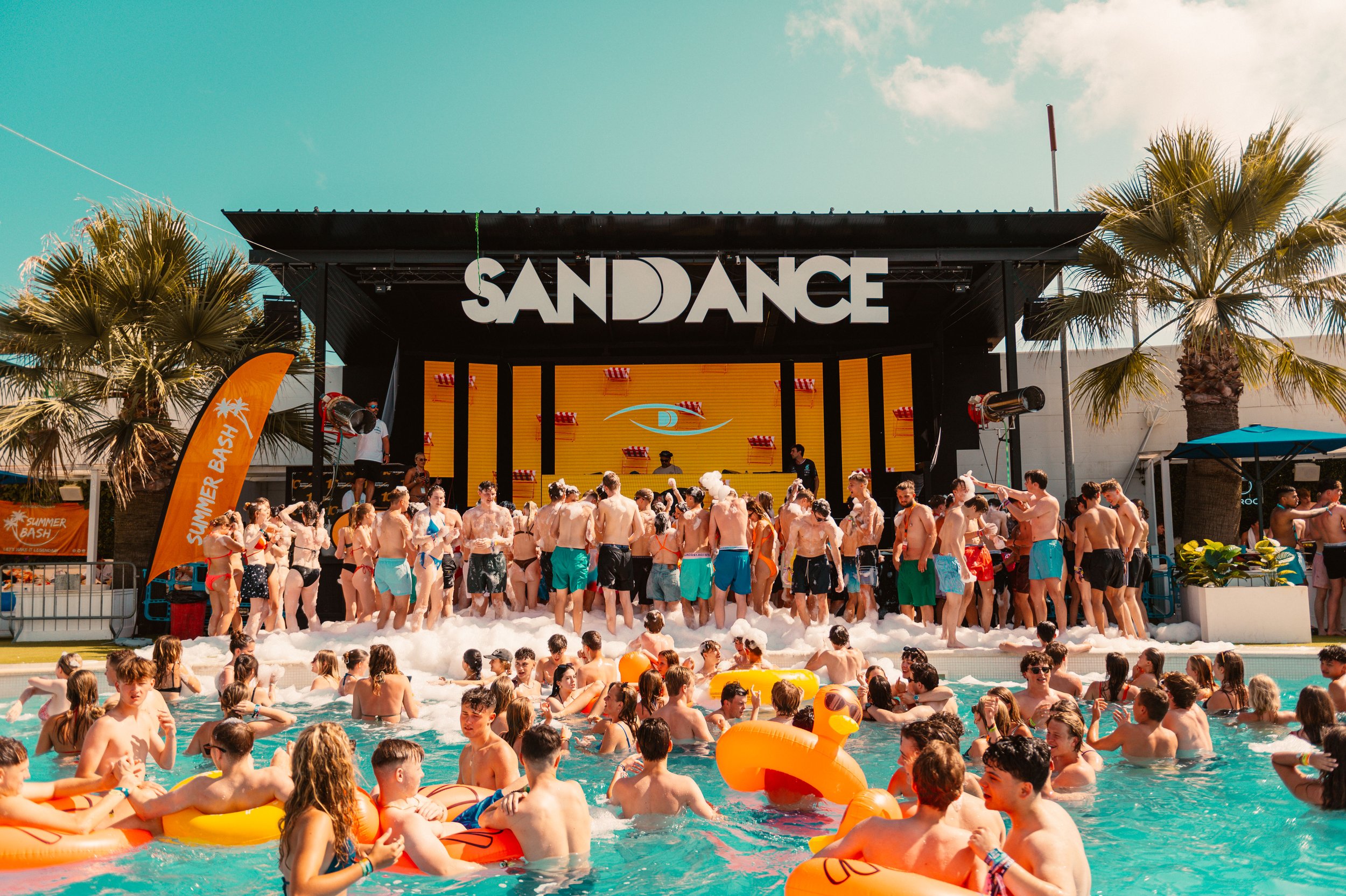 People enjoying a pool party with floaties and foam near a stage labeled 'Sandance,' under clear skies.