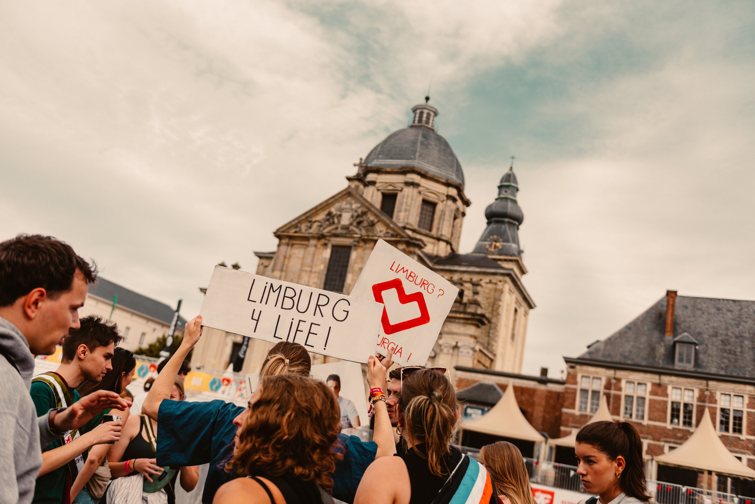 People at a public event holding signs saying 'Limburg 4 Life' and 'Limburg?' with a heart symbol, in front of a historic building with a dome and spire.