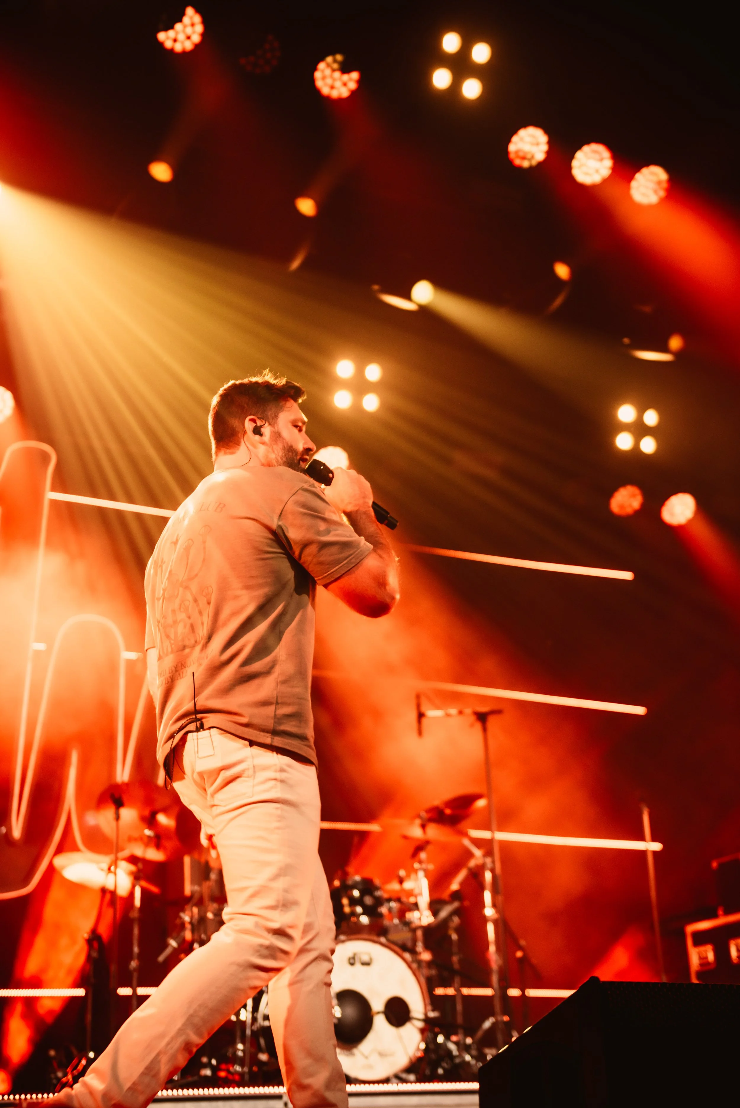 Male performer singing on stage with microphone under orange stage lights, drum set in background.