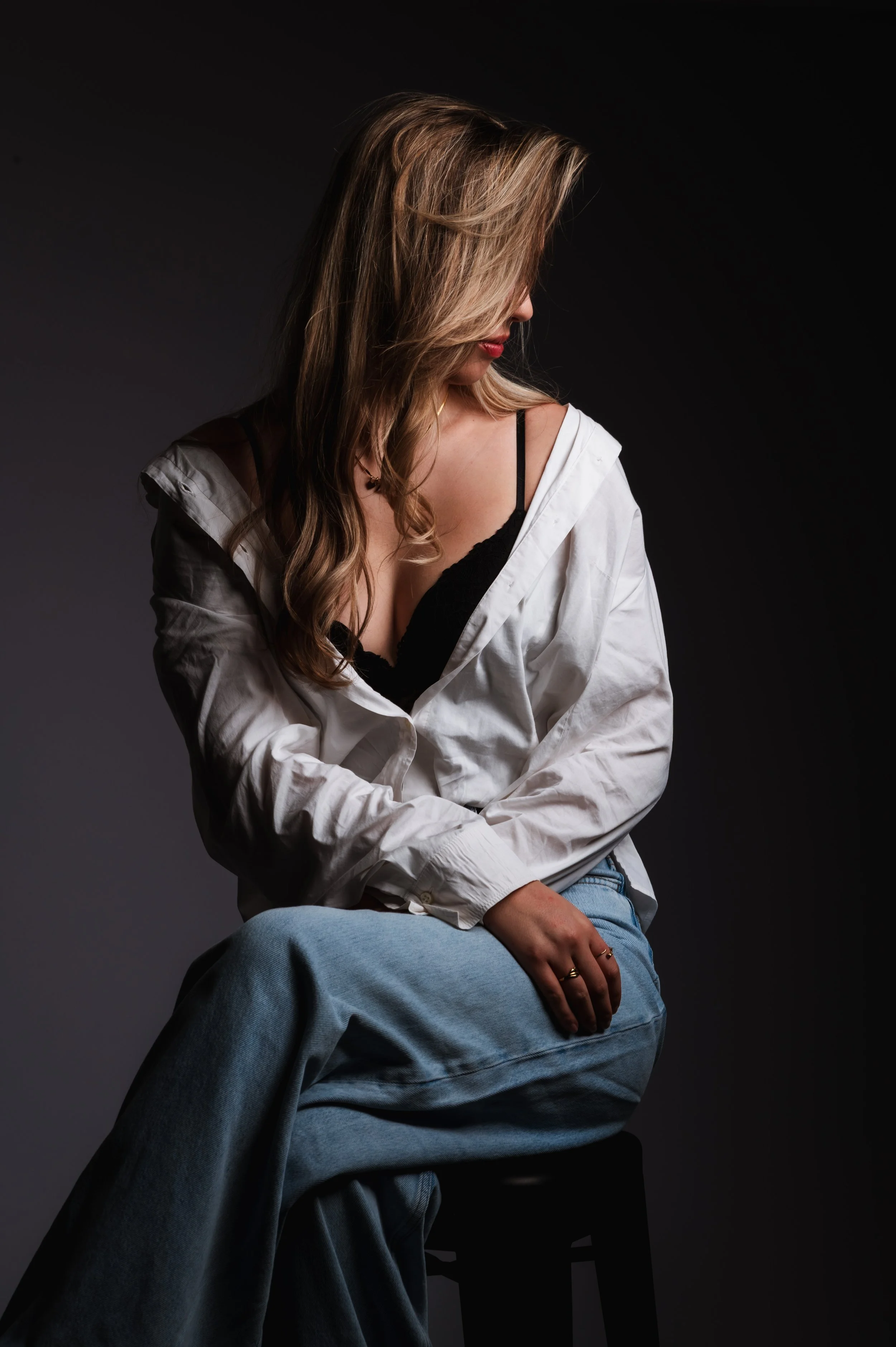 A woman with long, wavy hair, wearing a white shirt draped off her shoulders, a black lace bralette, and light blue jeans, sitting on a black stool against a dark background, looking downward.