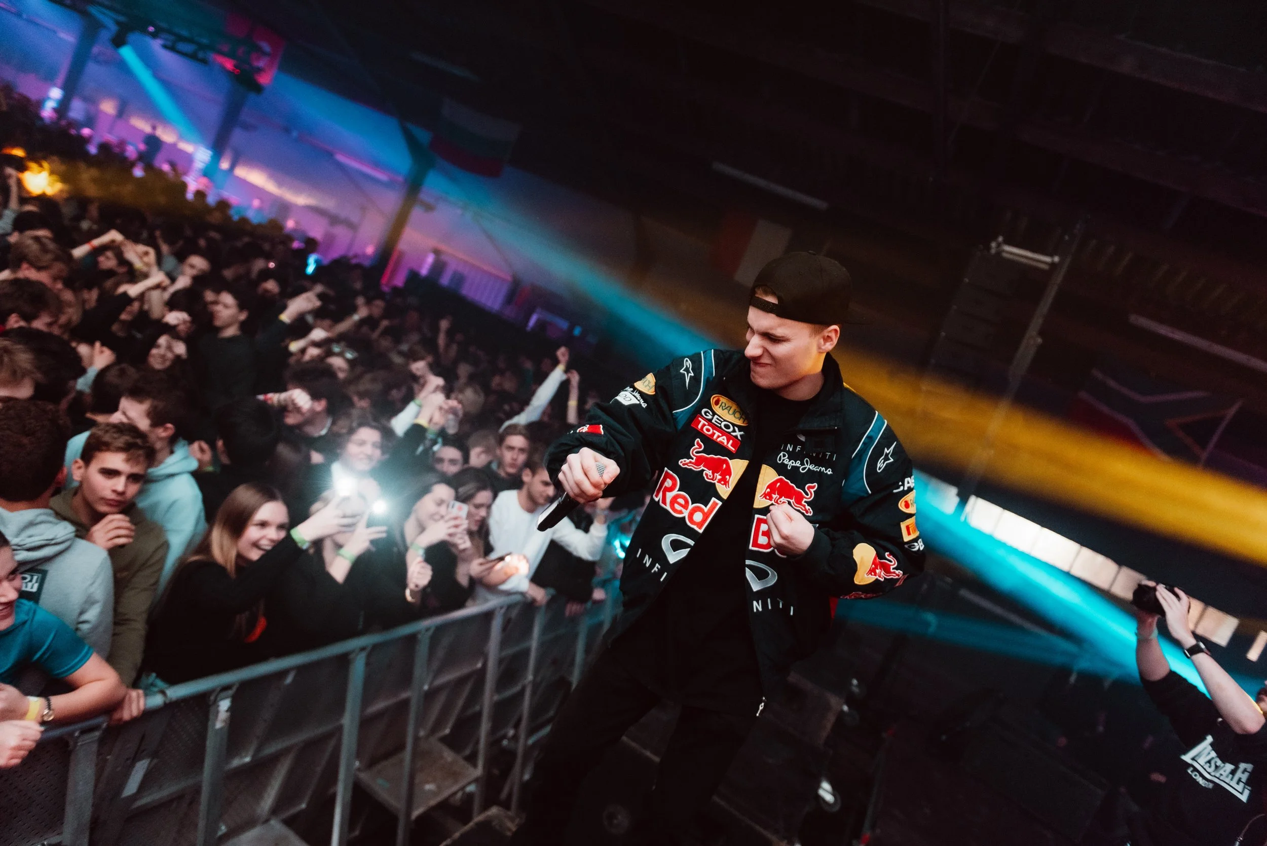 Performer in a Red Bull jacket on stage, interacting with a large, energetic crowd at a concert or event, with colorful lights and people taking photos.