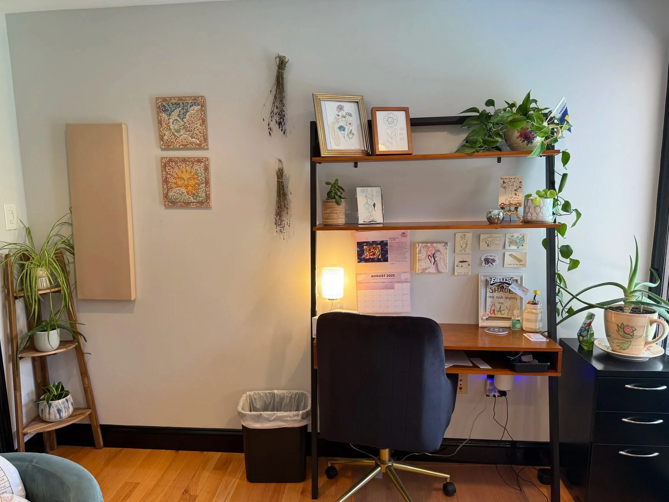 Home office setup with a black desk, black swivel chair, shelves with framed artwork and plants, wall decorations, a small black dresser, and various decorative items and plants.