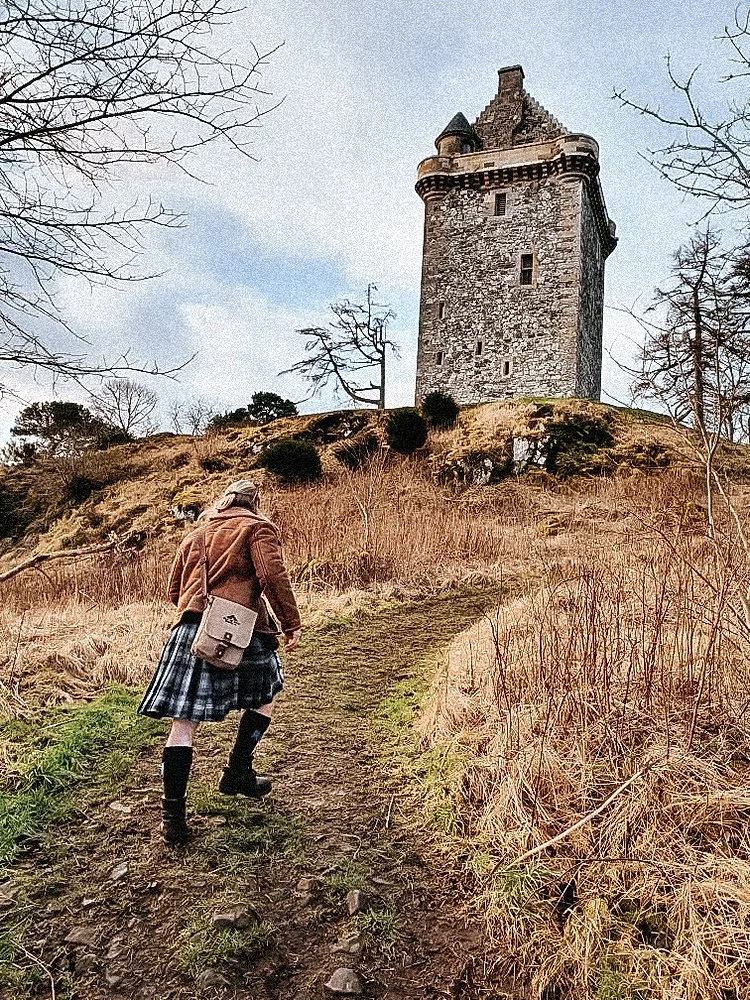A person in a brown jacket and plaid skirt walking up a dirt trail toward a tall, historic stone tower on a hill, with leafless trees around.