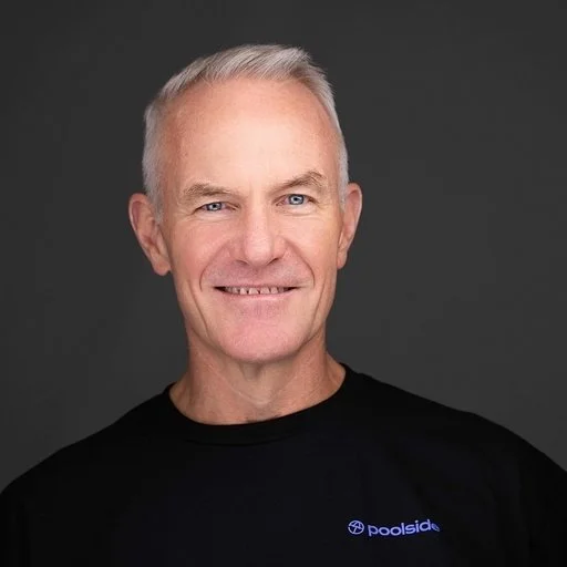 A middle-aged man with short gray hair, smiling, wearing a black T-shirt with a small logo on the chest, against a plain dark background.